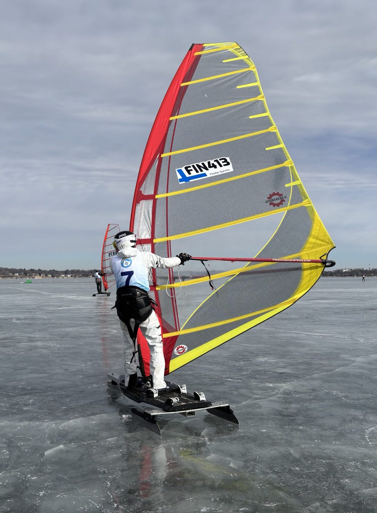 Person ice sailing on a frozen lake with a yellow and red sail under a cloudy sky. Other ice sailors are visible in the distance.