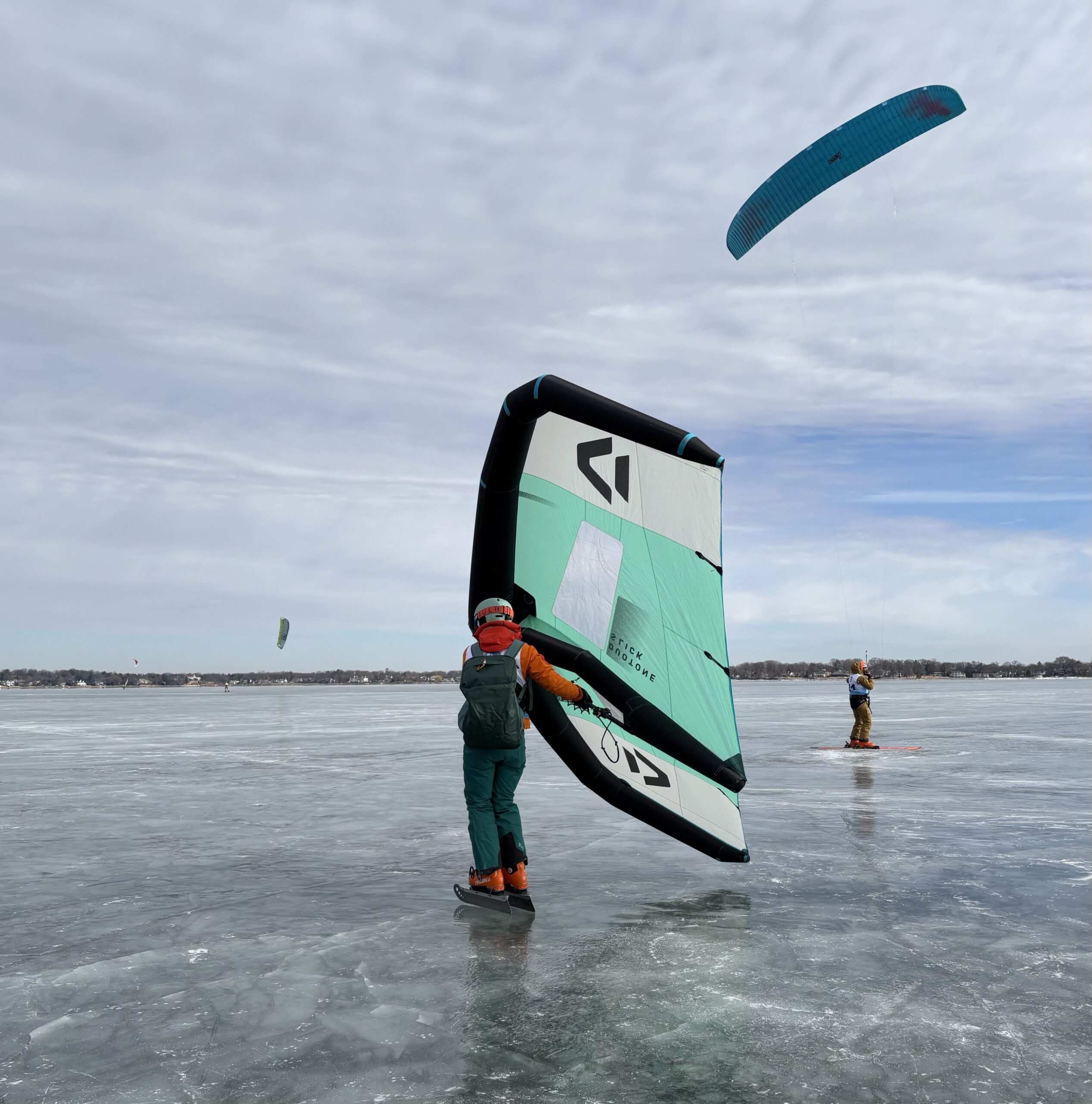 Two people kiteboarding on a frozen lake, each holding a kite wing. The sky is cloudy, and the ice is visible beneath them.