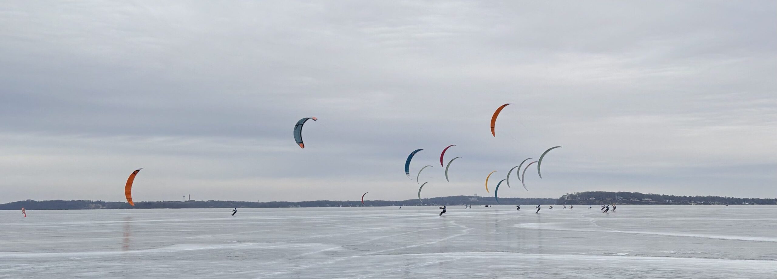 Several people kitesurf on a large, frozen body of water. Colorful kites are visible against a cloudy sky, and trees line the distant shore.