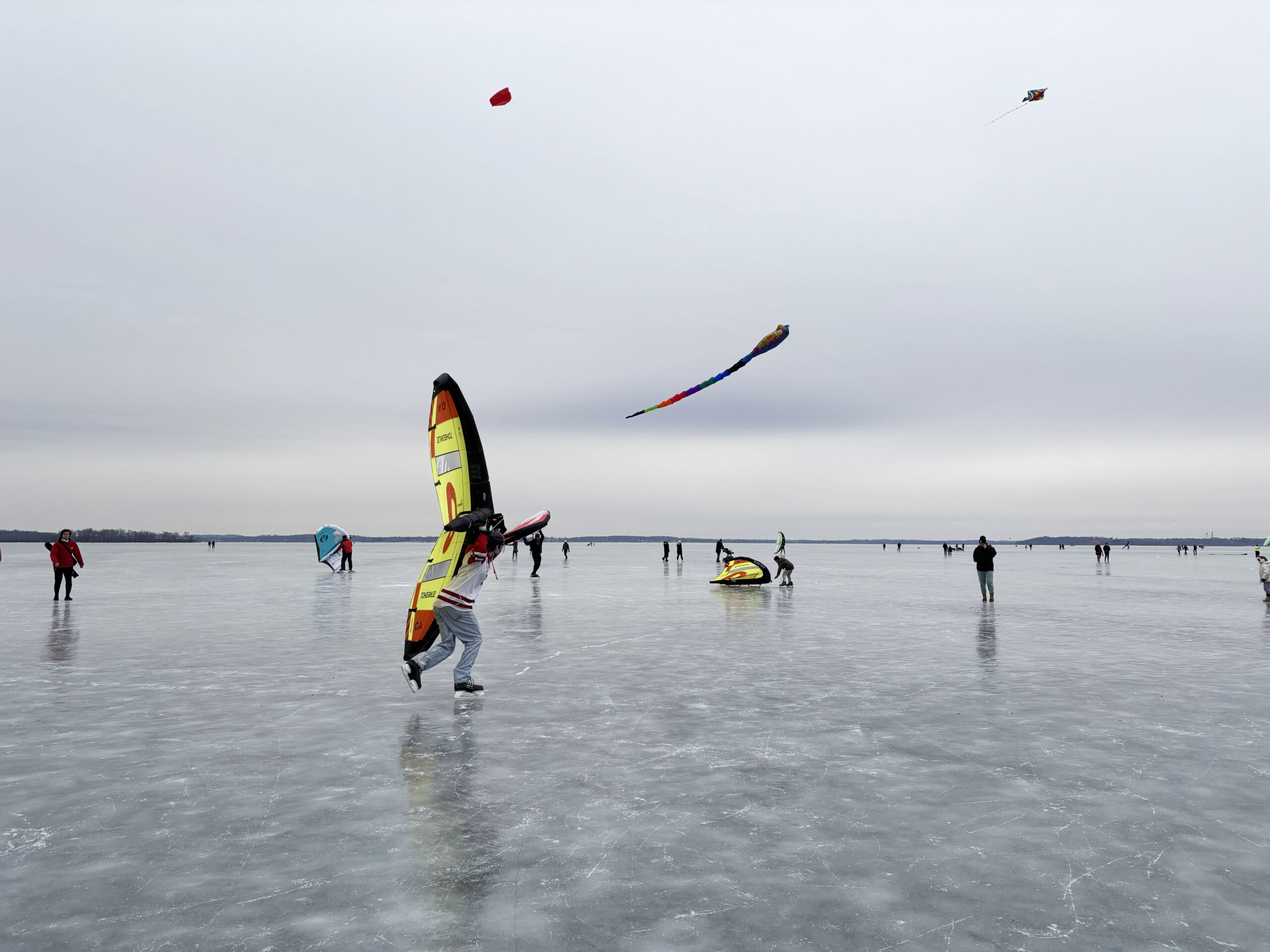 People participating in ice kiting on a frozen lake, with several colorful sails and kites visible against a gray sky. The surface of the ice is clear and reflective.