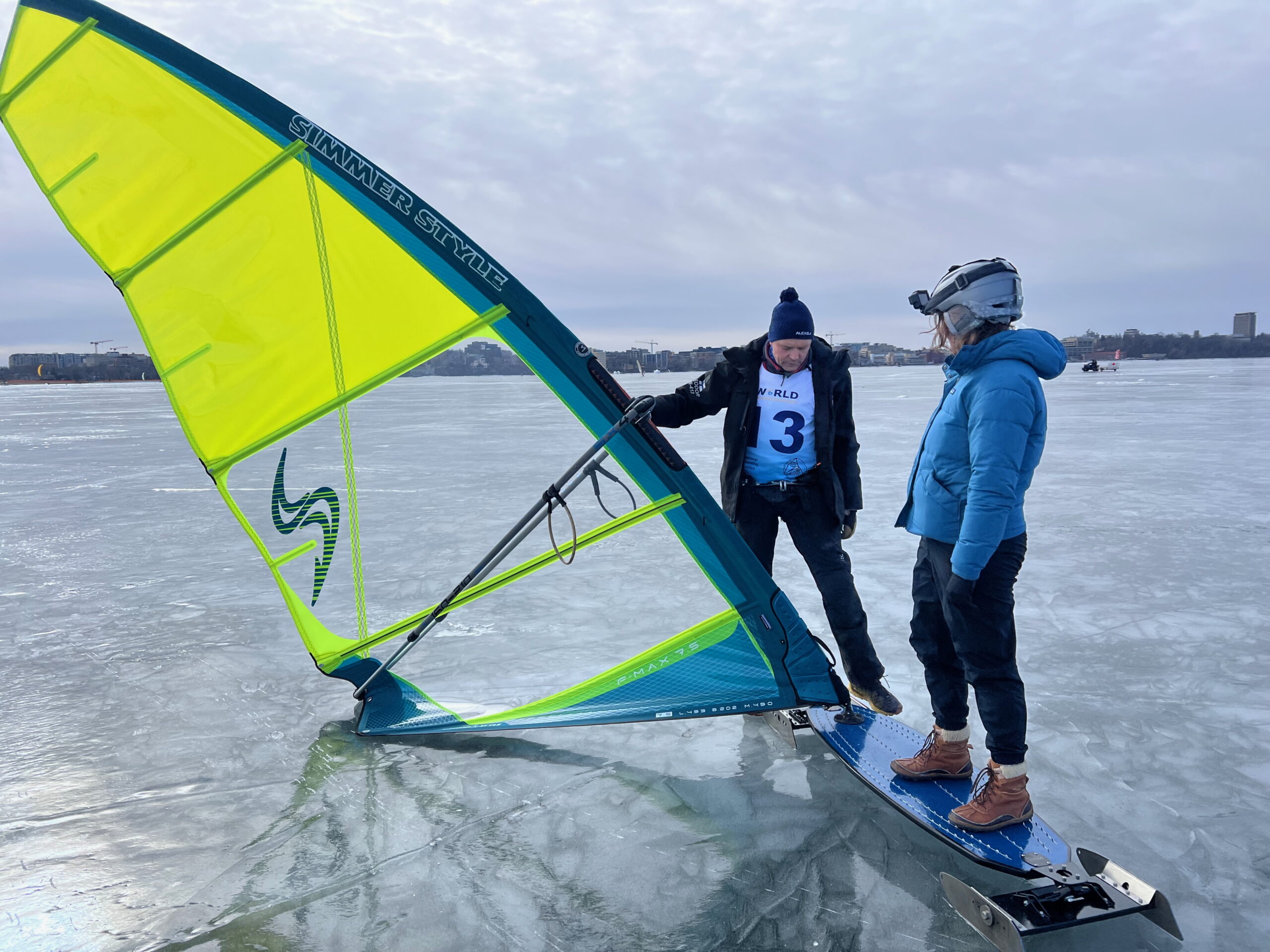 Two people stand on an ice-covered lake with a yellow and green ice sailing rig. One person wears a numbered vest and the other is in a blue jacket and helmet.