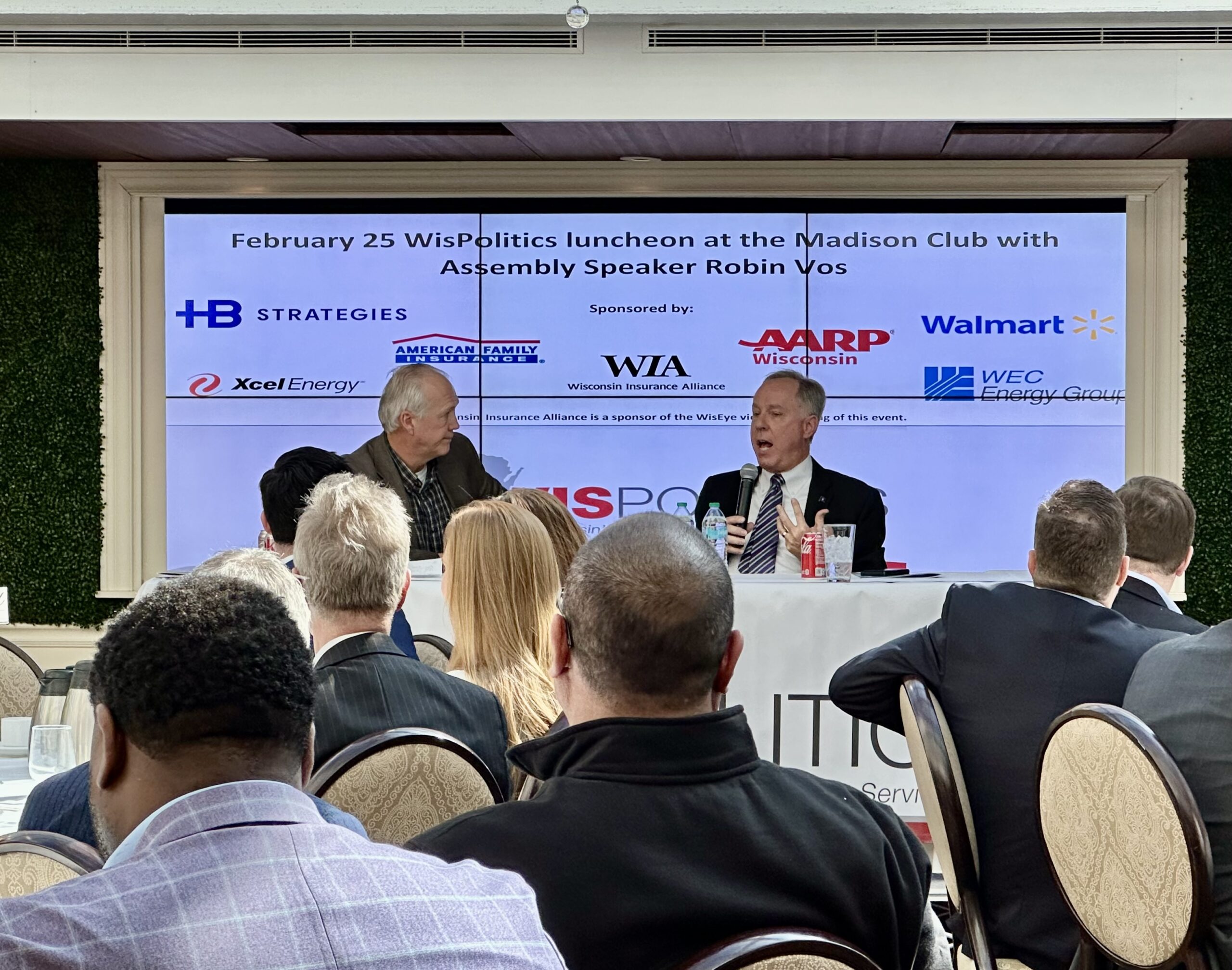 Two people sitting at a table with microphones, speaking at the WisPolitics luncheon. Banners and sponsorship logos are displayed behind them. Audience members are visible in the foreground.