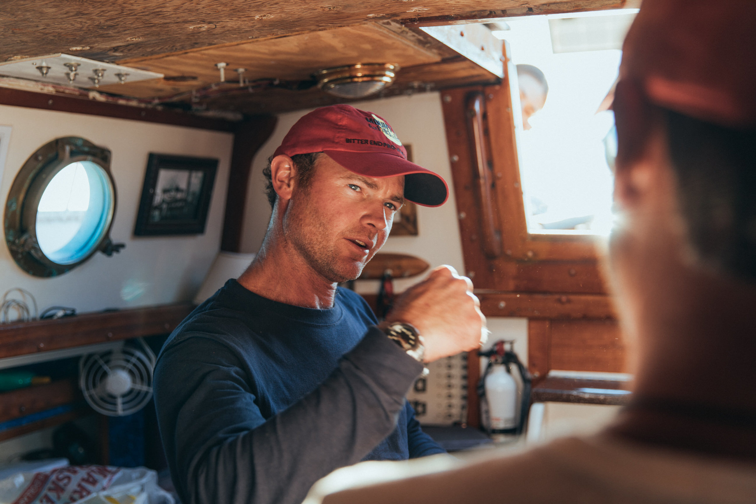 Man in a cabin with wooden interior gestures while talking to another person. He wears a red cap and navy shirt.