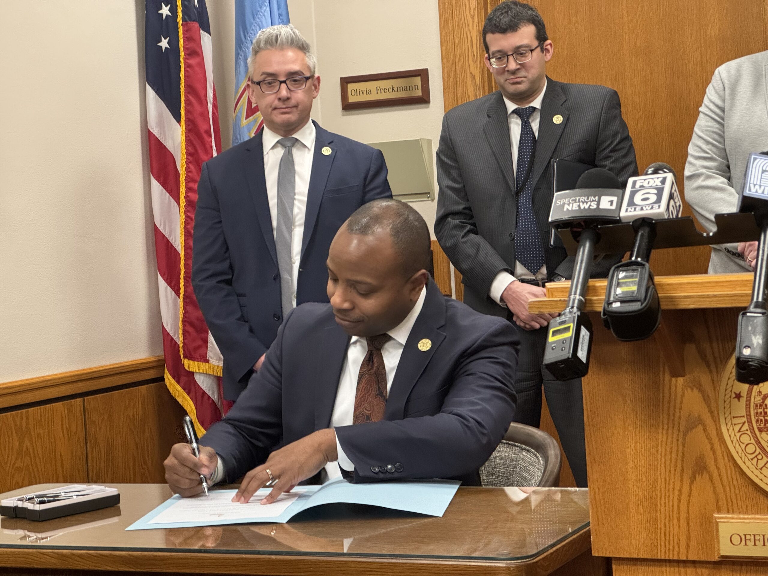 A man in a suit signs a document at a desk with microphones. Two other men stand behind him, observing. An American flag is partially visible in the background.