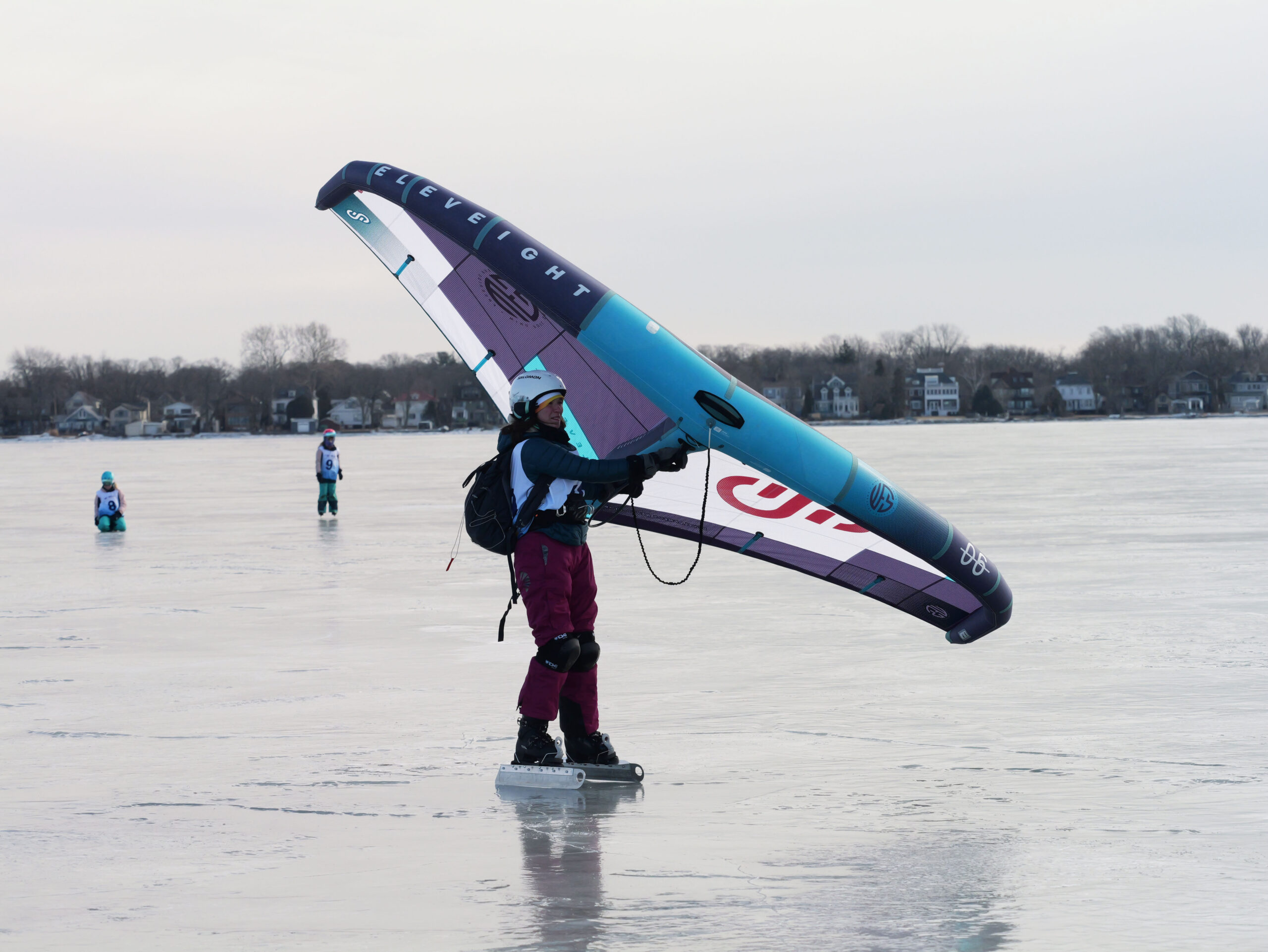 Person ice skating on a frozen lake while holding a large wing-shaped sail, with houses visible in the background.