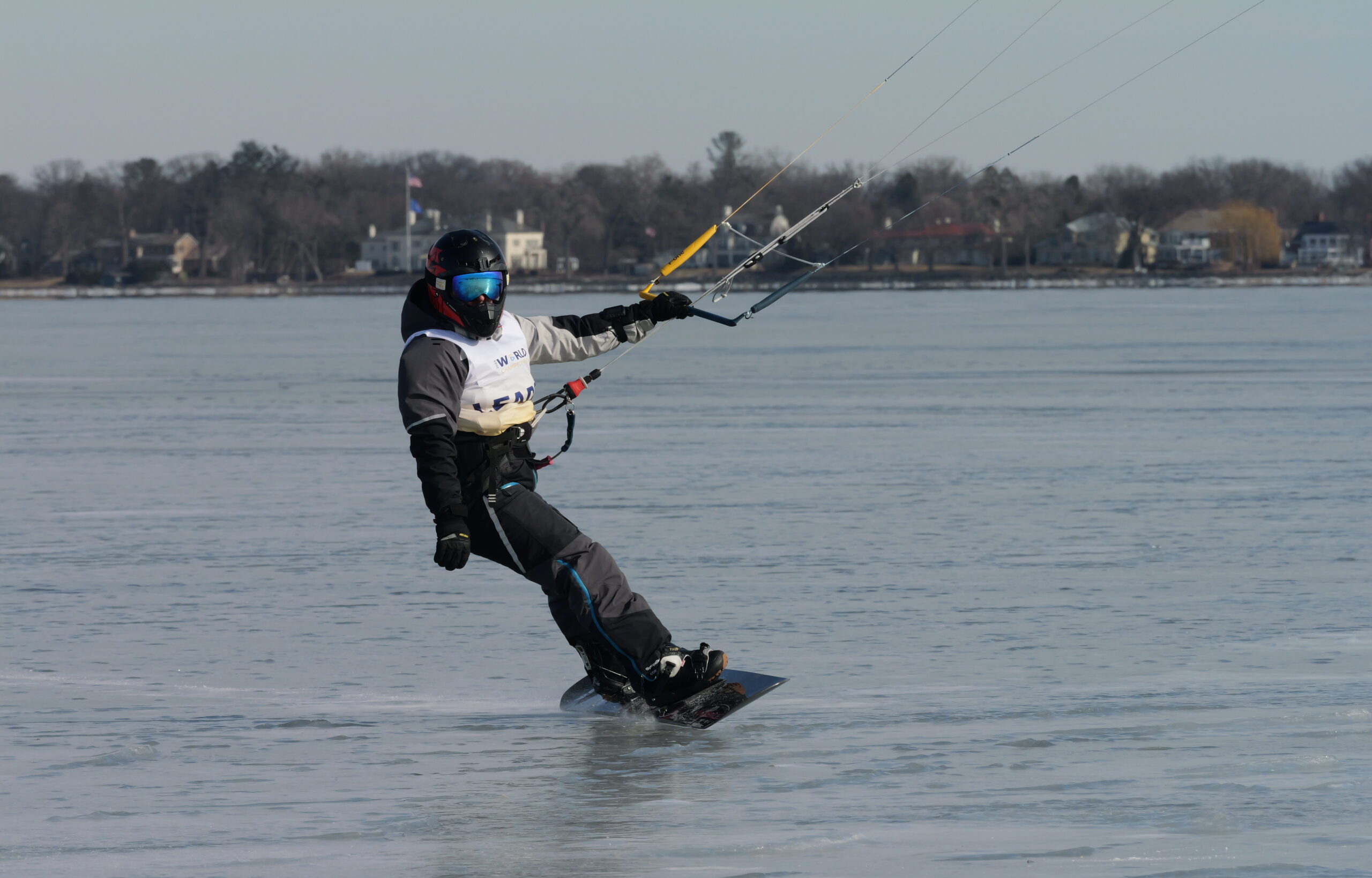 Person kitesurfing on a frozen lake, wearing a helmet and goggles. Trees and houses are visible in the background.