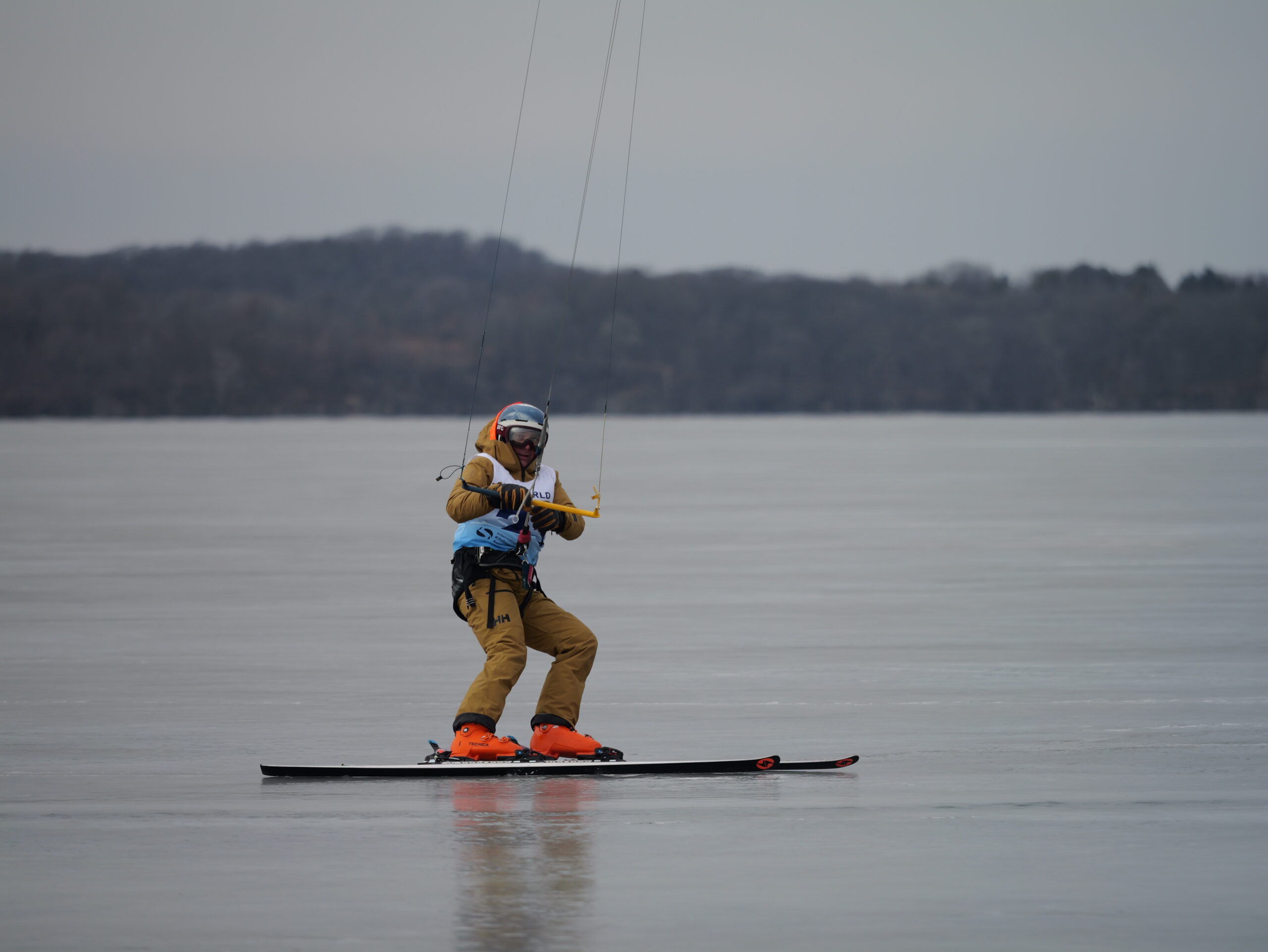 Person snowkiting on a frozen lake, wearing brown clothing and orange boots, holding onto a kite handle.