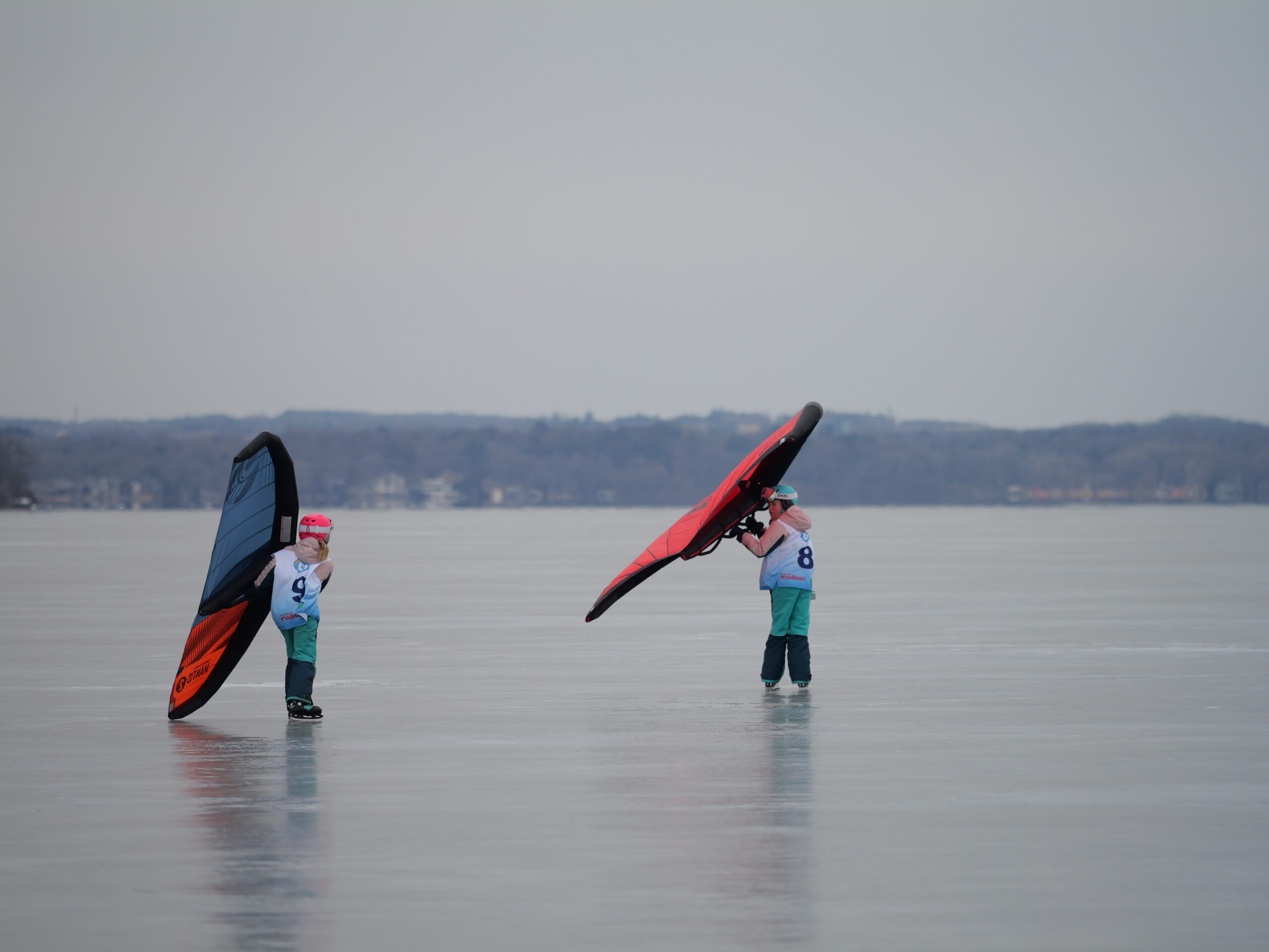 Two children wearing helmets hold large kiteboarding wings on an open ice-covered surface with a distant treeline in the background.