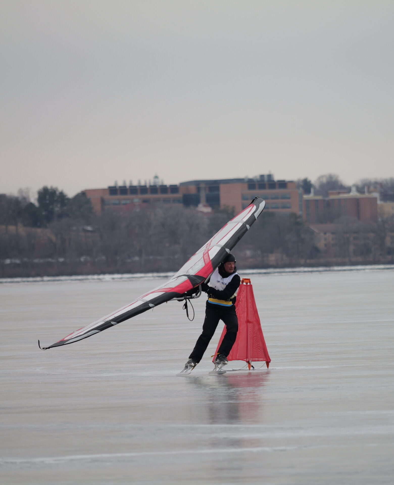 A person ice sailing on a frozen lake using a red and black windsurfing sail. Trees and buildings are visible in the background under an overcast sky.