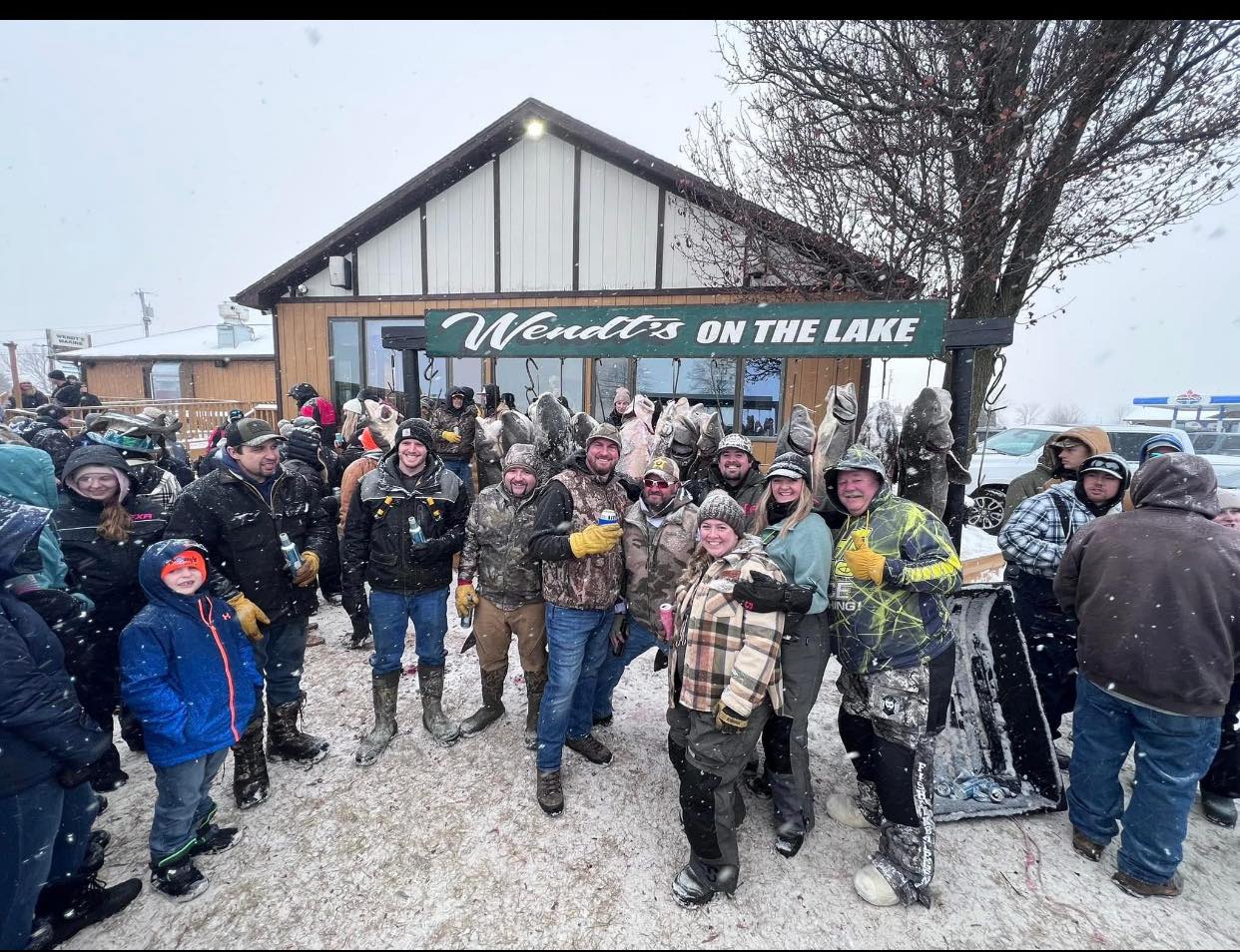 A group of people stand outside Wendts on the Lake in winter attire, holding fish and posing in a snowy environment.