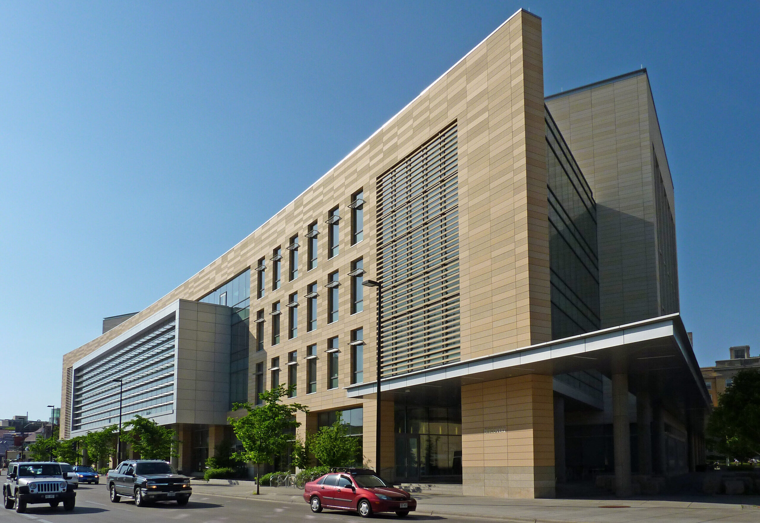 Modern beige building with large windows, surrounded by trees and parked cars, under a clear blue sky.