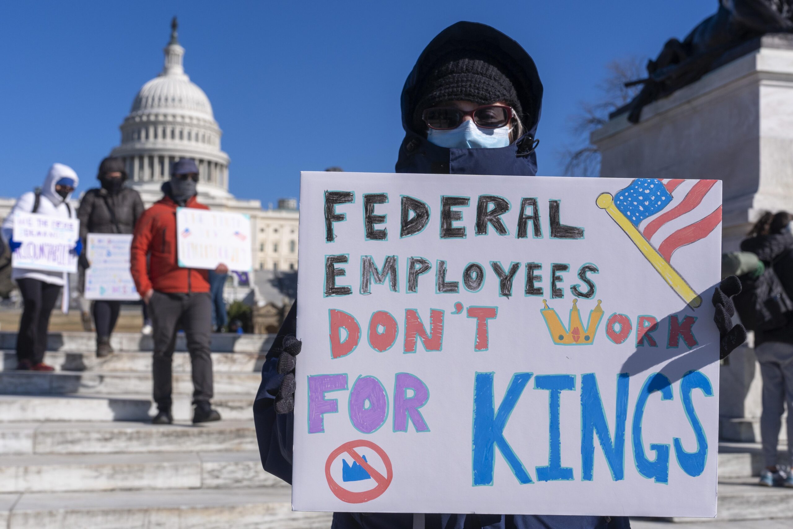 A person holds a sign reading Federal Employees Dont Work for Kings in front of the U.S. Capitol, with others holding signs in the background.