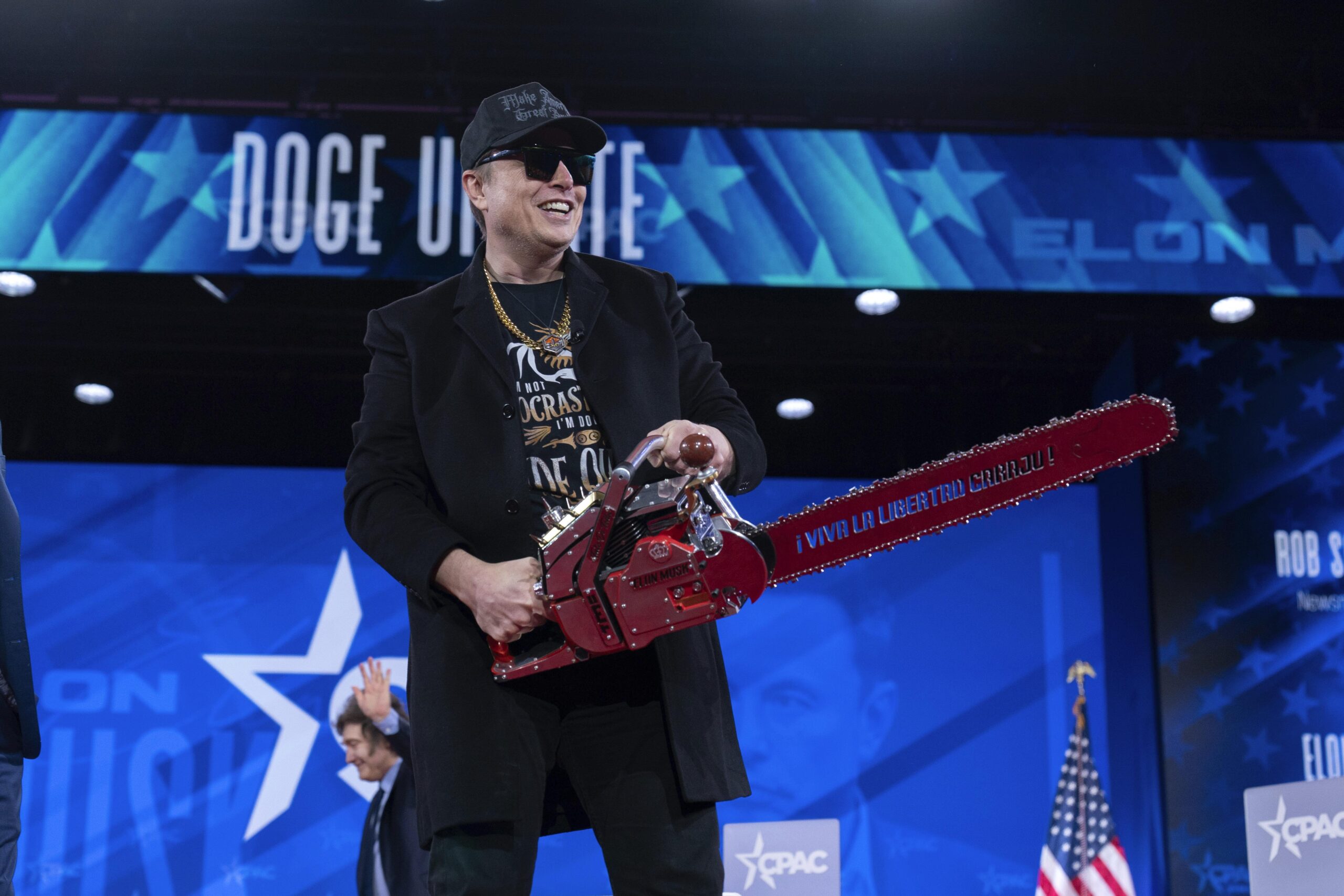 Elon Musk wearing sunglasses and a cap holds a large decorative chainsaw on stage, with a backdrop displaying Doge Unite.