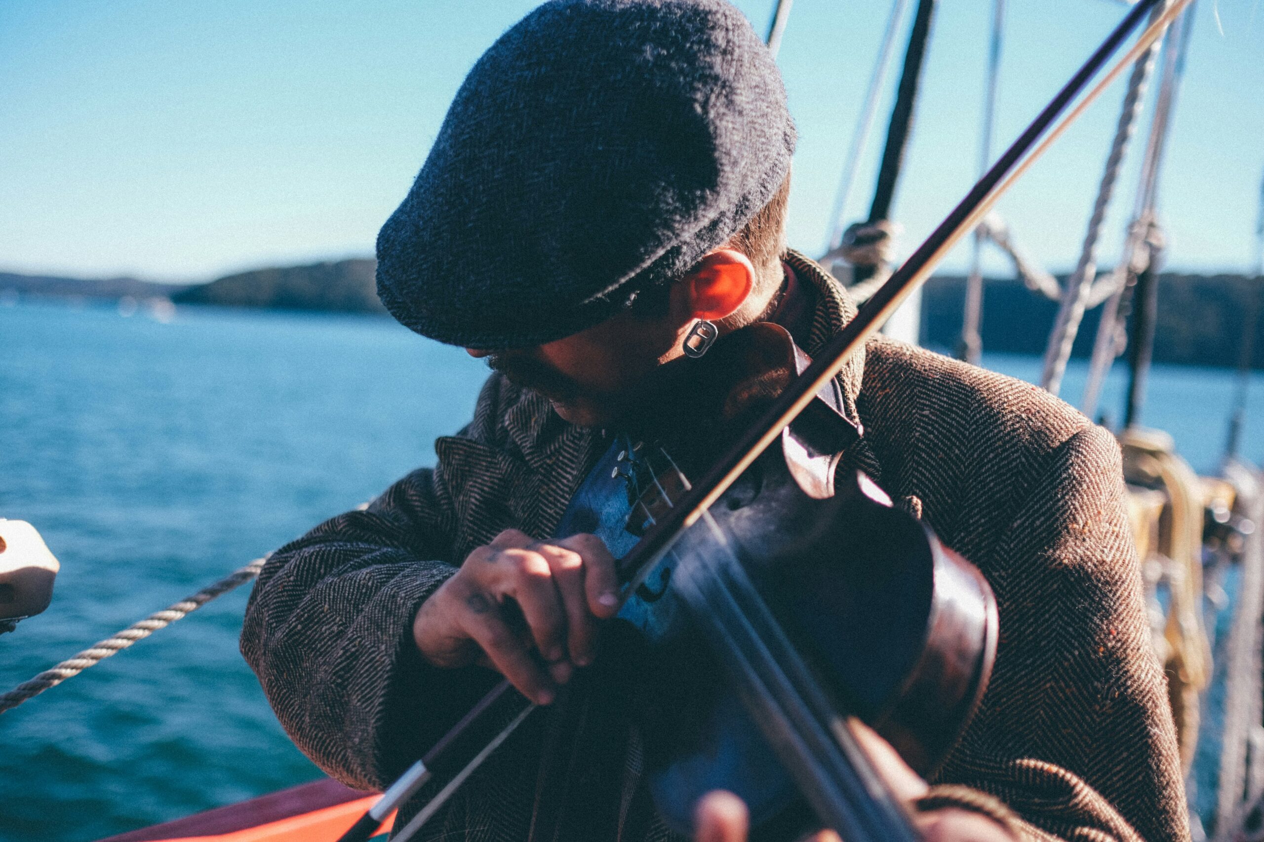 A person in a cap and jacket plays the violin on a boat with a view of the sea and distant shoreline.