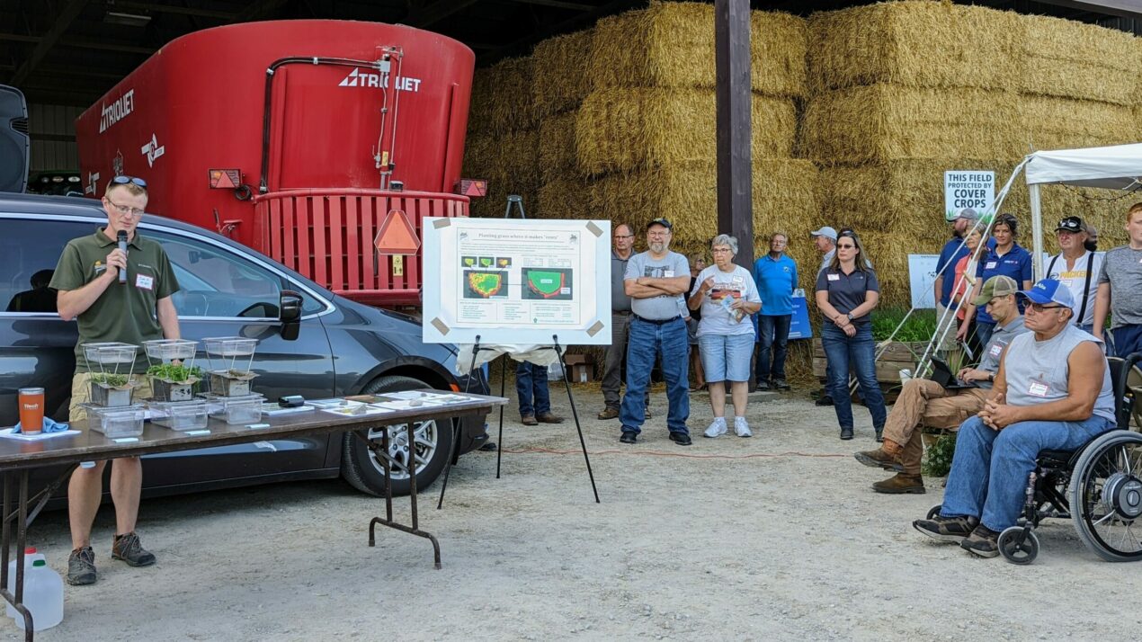 A man speaks into a microphone at an outdoor event with a poster board and plants on a table. People are seated and standing, listening. Hay bales and machinery are in the background.