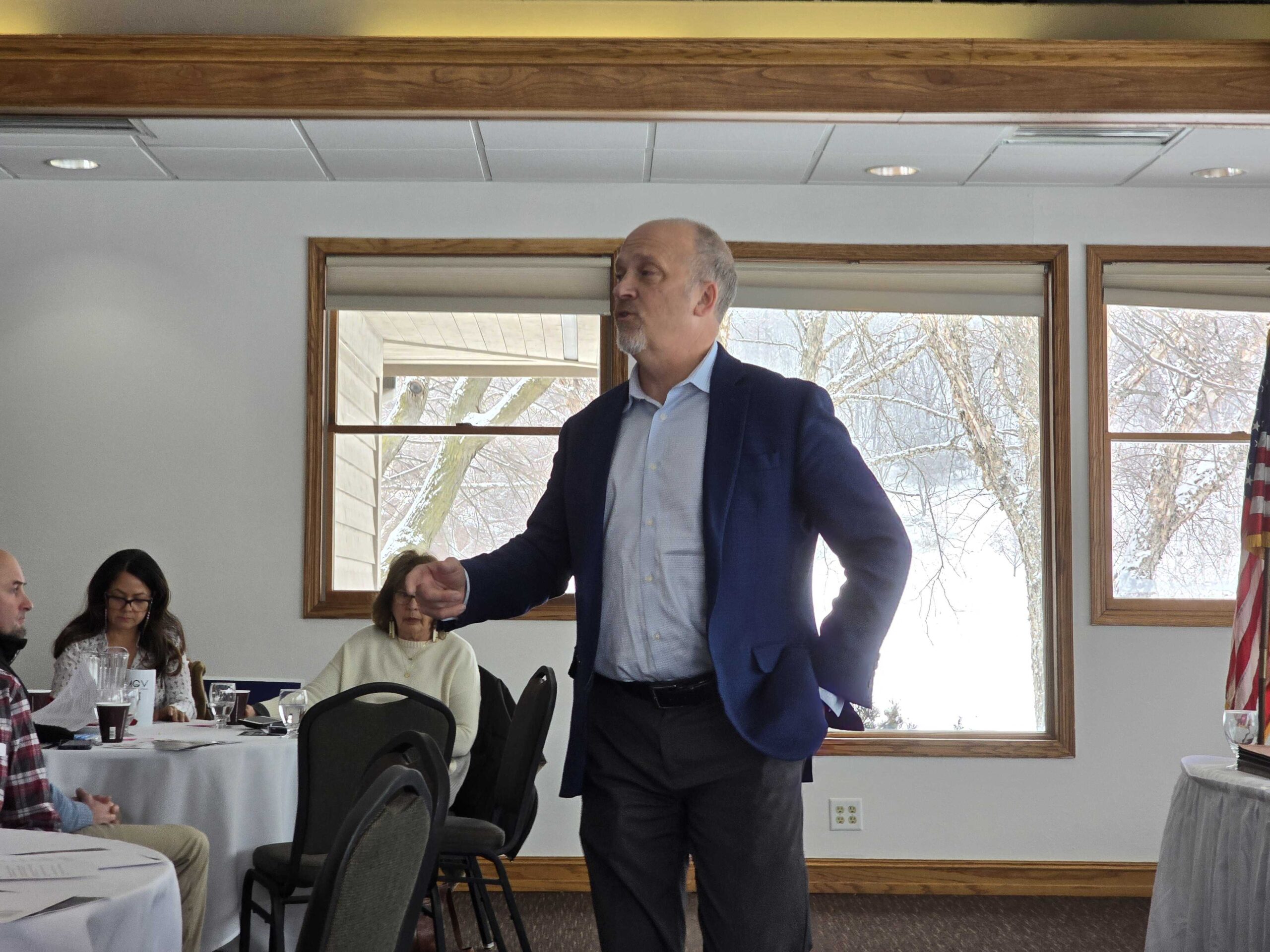 Man in a suit stands and speaks in a room with round tables and an audience, next to a window showing a snowy landscape.