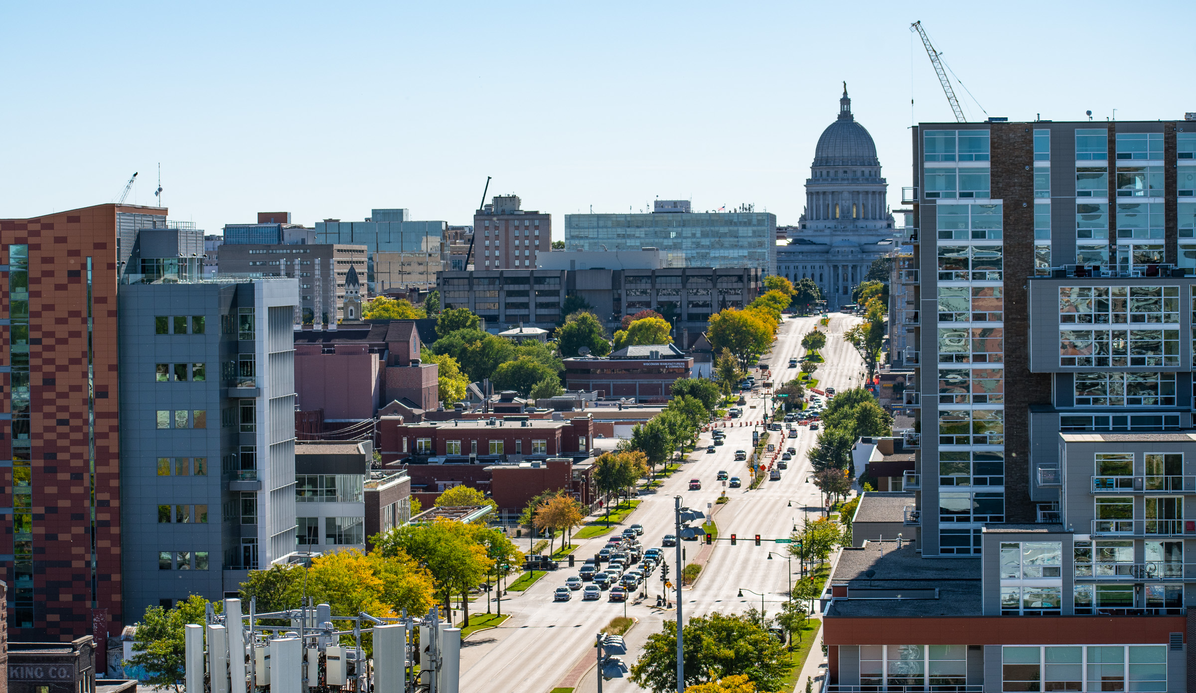 A busy city street lined with trees and buildings leads to a distant dome-shaped capitol building under a clear blue sky.