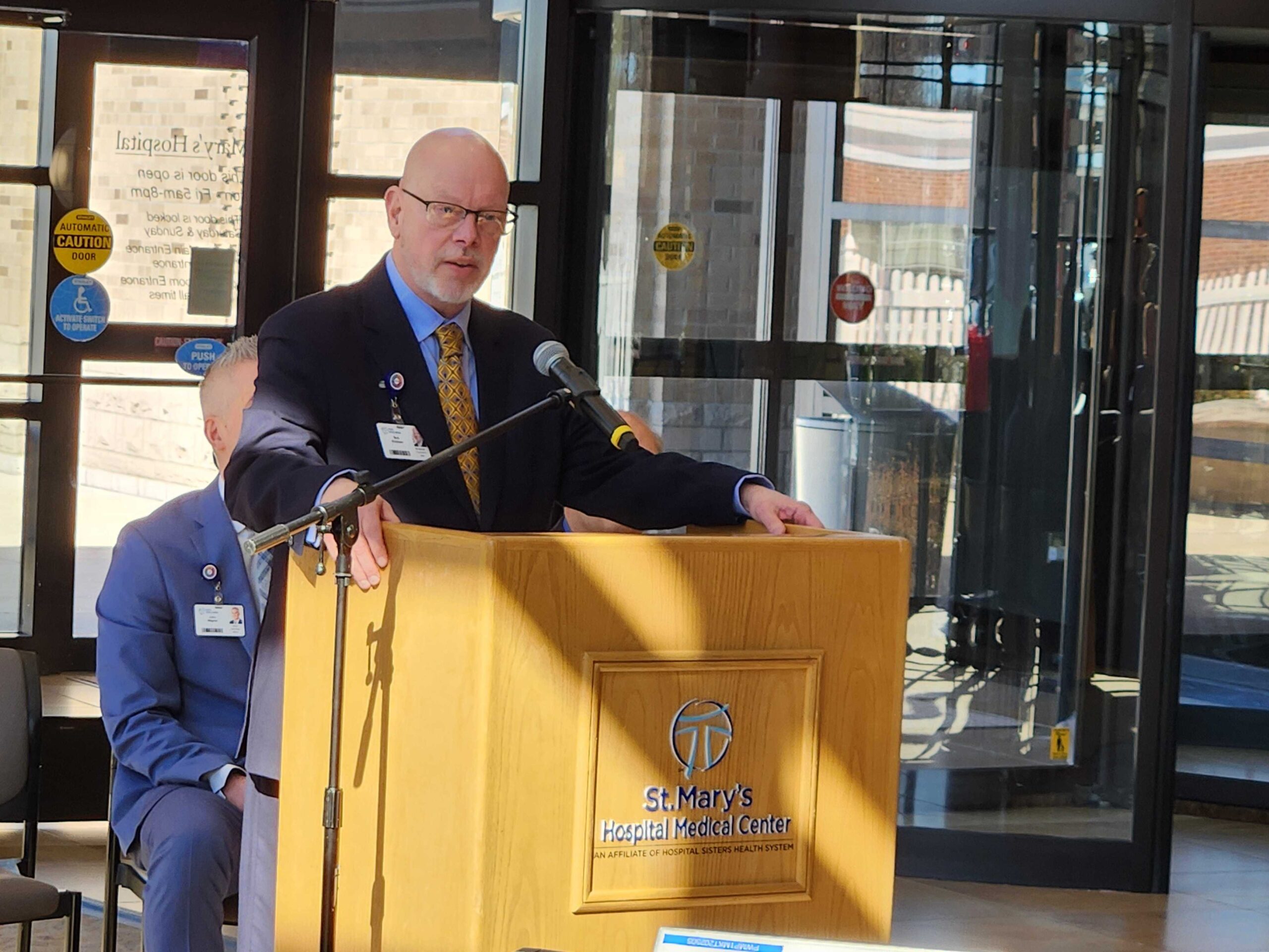 A man in a suit speaks at a podium labeled St. Marys Hospital Medical Center with a microphone in front, inside a building with glass doors in the background.