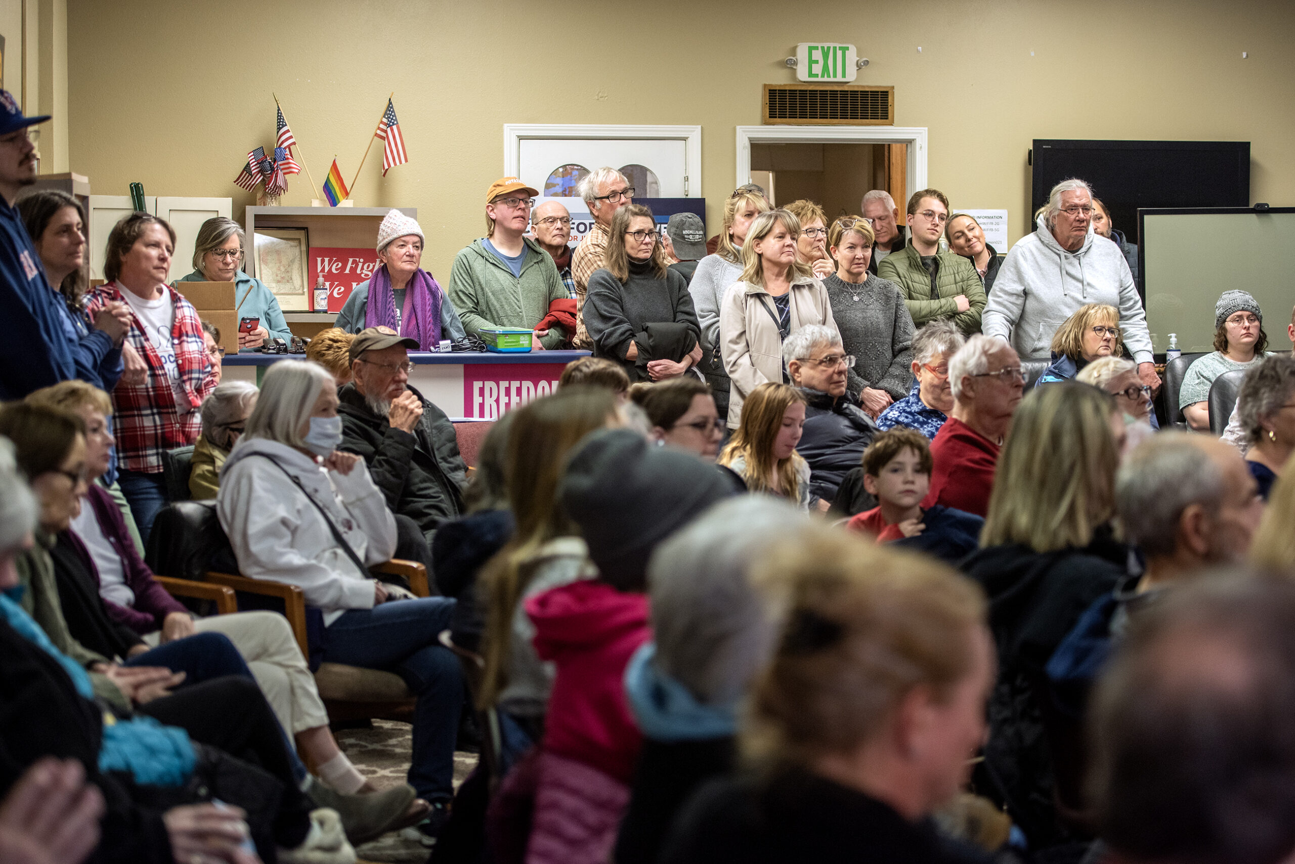 A group of people sit and stand in a room, attentively watching something. There are flags and a sign that reads Freedom in the background.