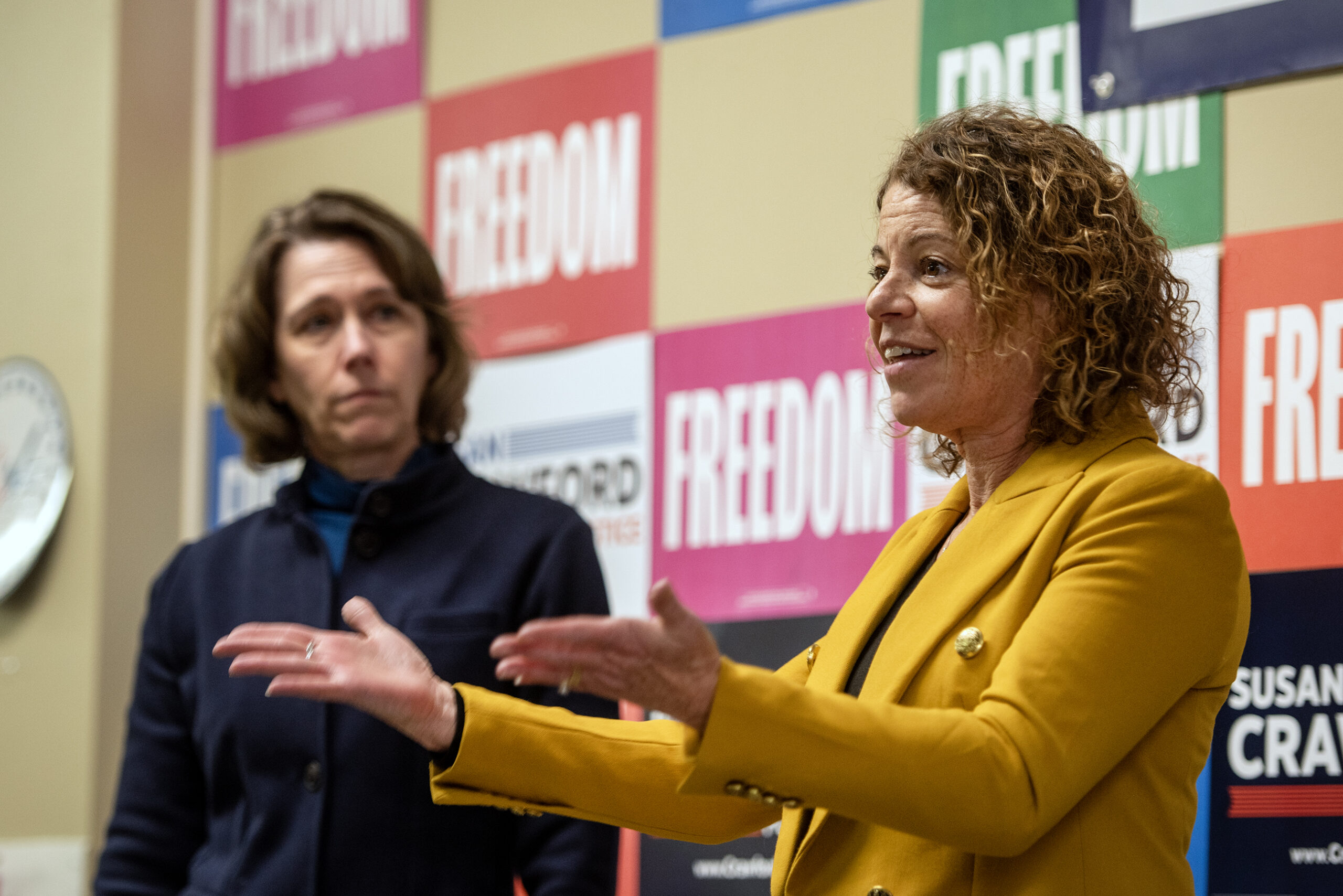 Two people stand in front of a wall with FREEDOM signs. The person on the right gestures while speaking, wearing a yellow jacket.