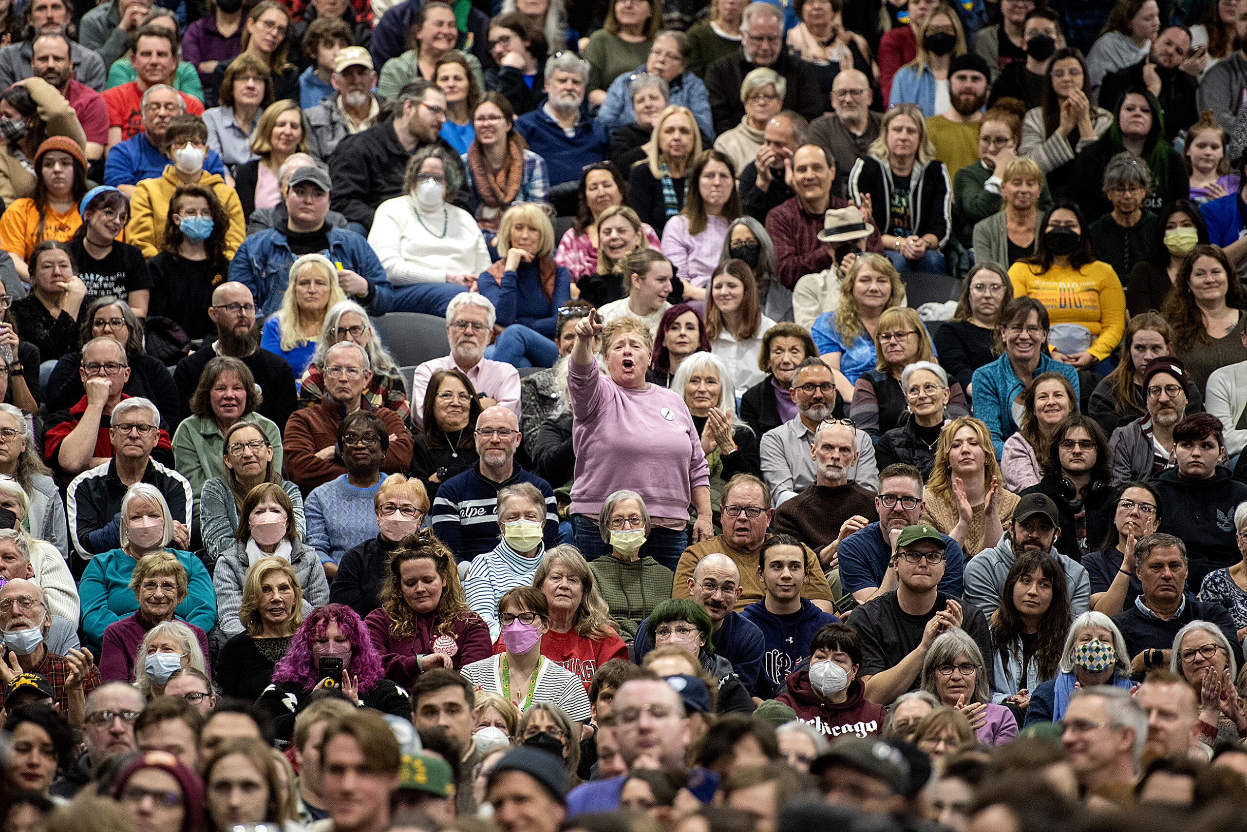 A large, diverse crowd sits closely packed in an indoor venue. Some individuals wear face masks. People are attentively looking ahead, with varied expressions and postures.