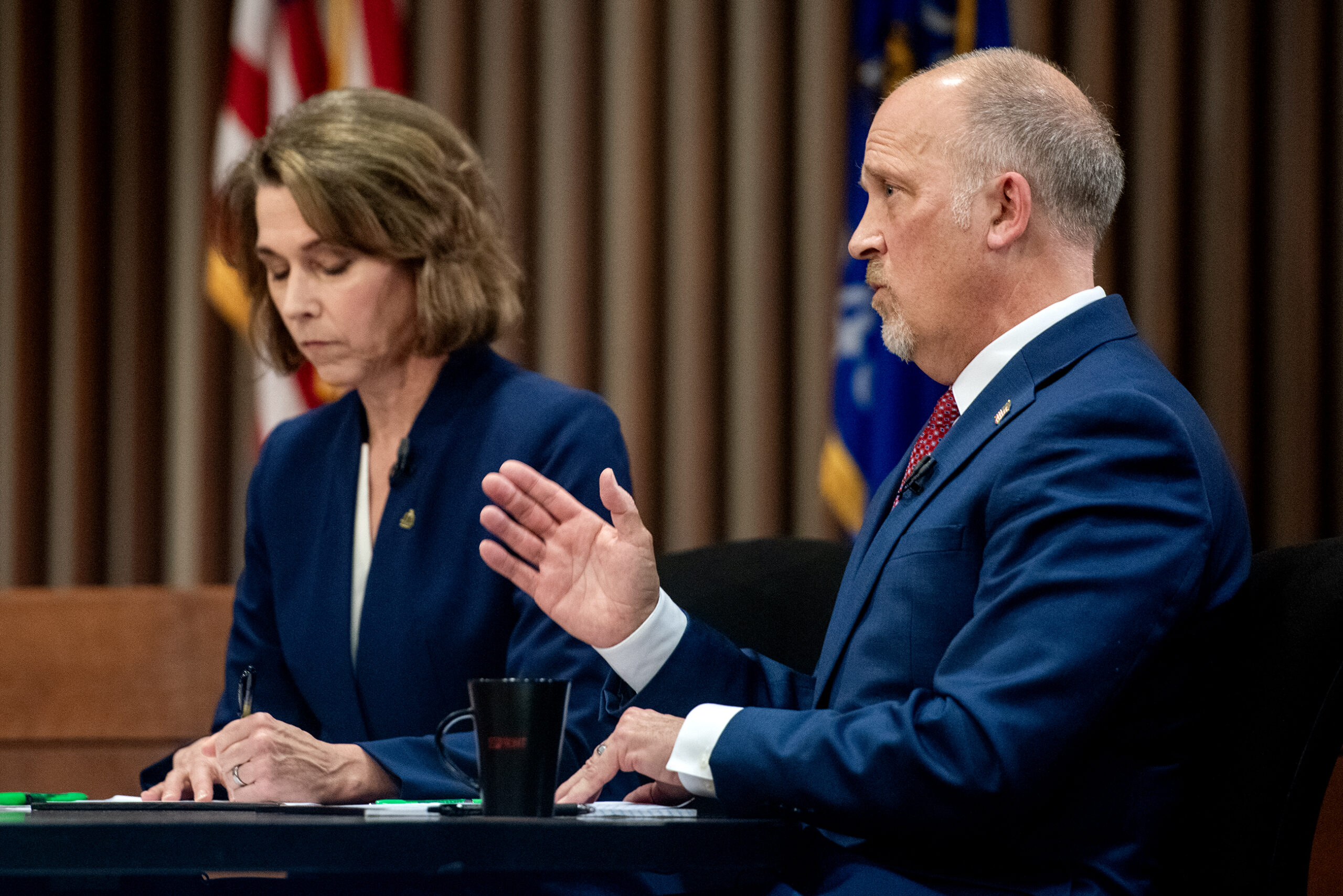 Two people in formal attire are seated at a table. One is speaking with a hand gesture while the other writes notes. Background includes flags and vertical blinds.