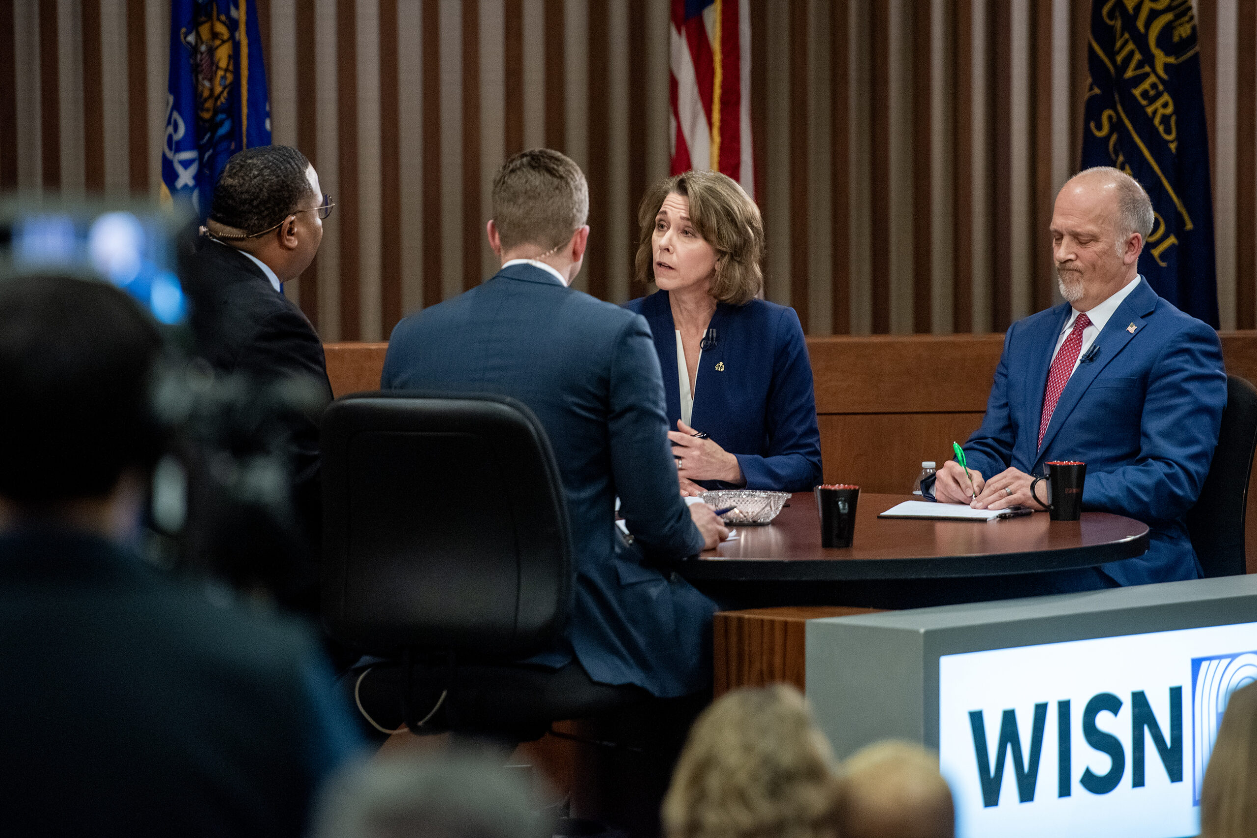 Three people seated at a round table engaged in a discussion, with a moderator nearby. A WISN branded desk is visible in the foreground.