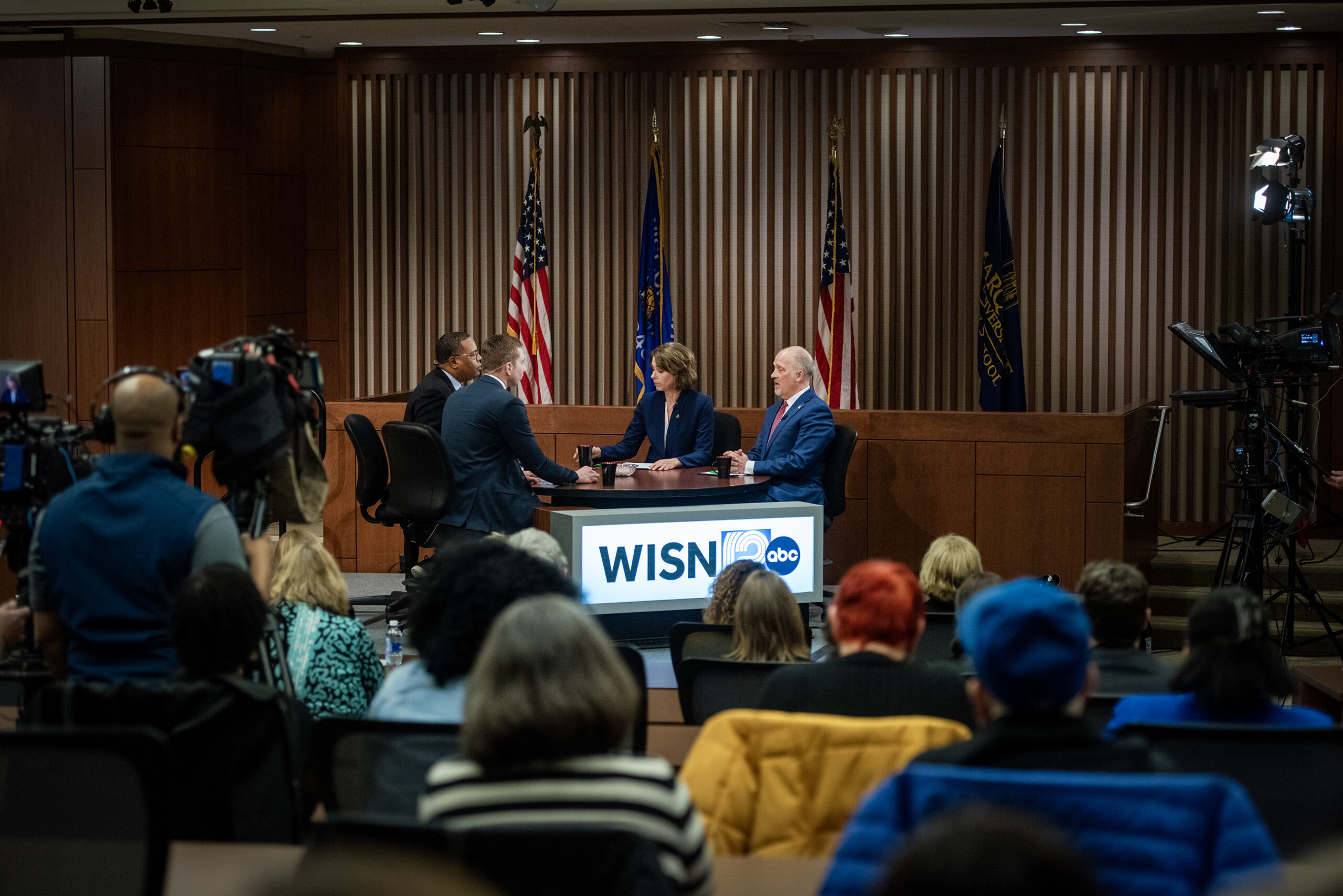 Four people seated at a news debate table with WISN 12 ABC branding, surrounded by cameras and audience members. American flags are visible in the background.