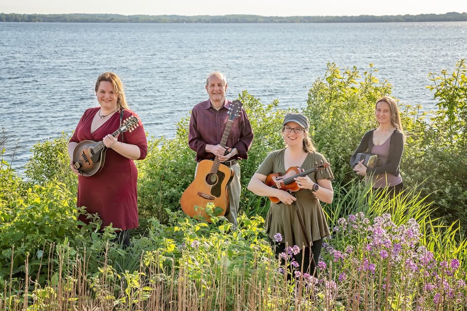 Four people holding musical instruments stand by a lakeside with greenery and flowers in the foreground.