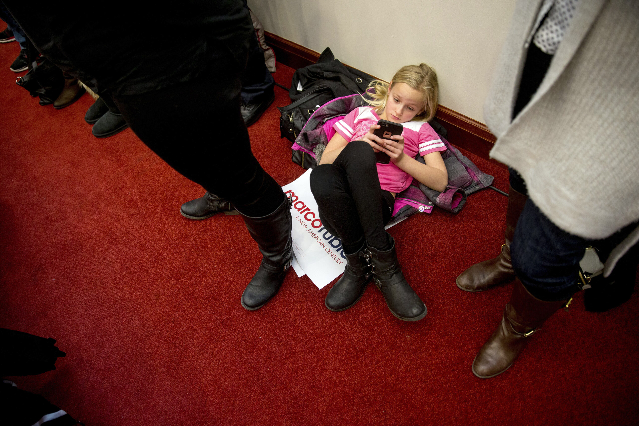 A girl in a pink shirt lays on the floor looking at a smart phone as adults stand around her