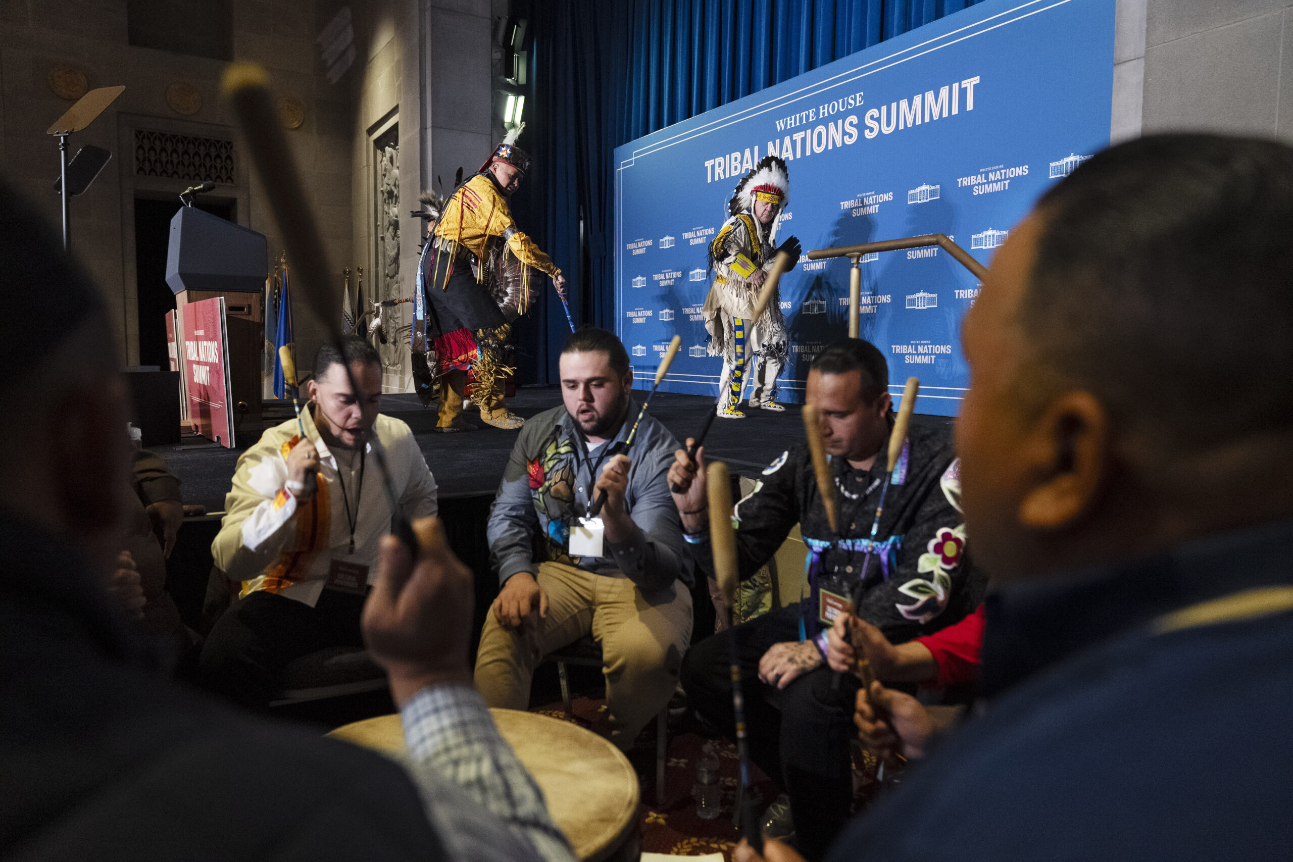 A group of people are dressed in traditional clothing, sitting around a drum, performing at the Tribal Nations Summit. Two individuals in regalia stand in front of a blue backdrop.