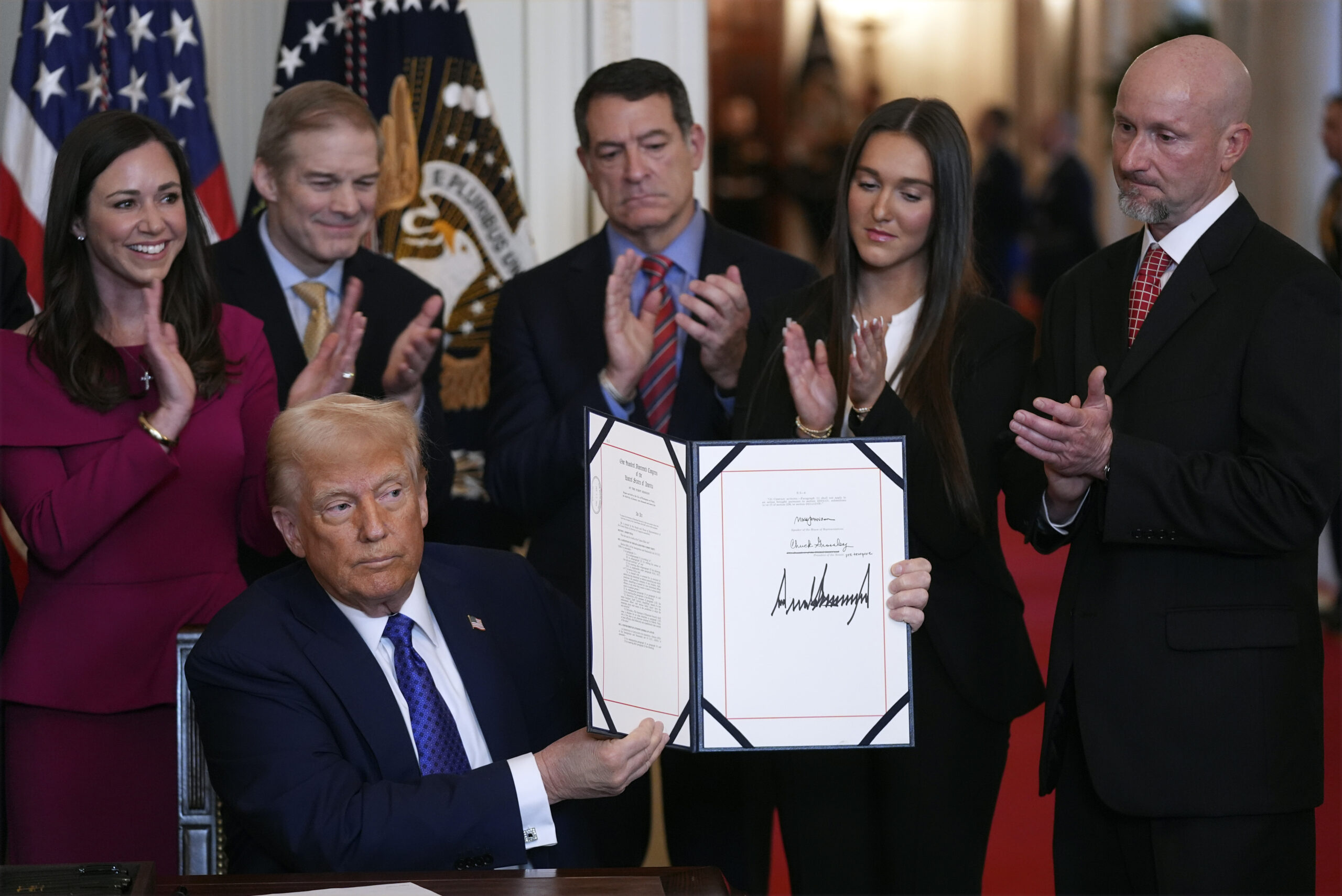 A man seated at a desk holds up a signed document, surrounded by five people applauding. An emblem and U.S. flags are in the background.