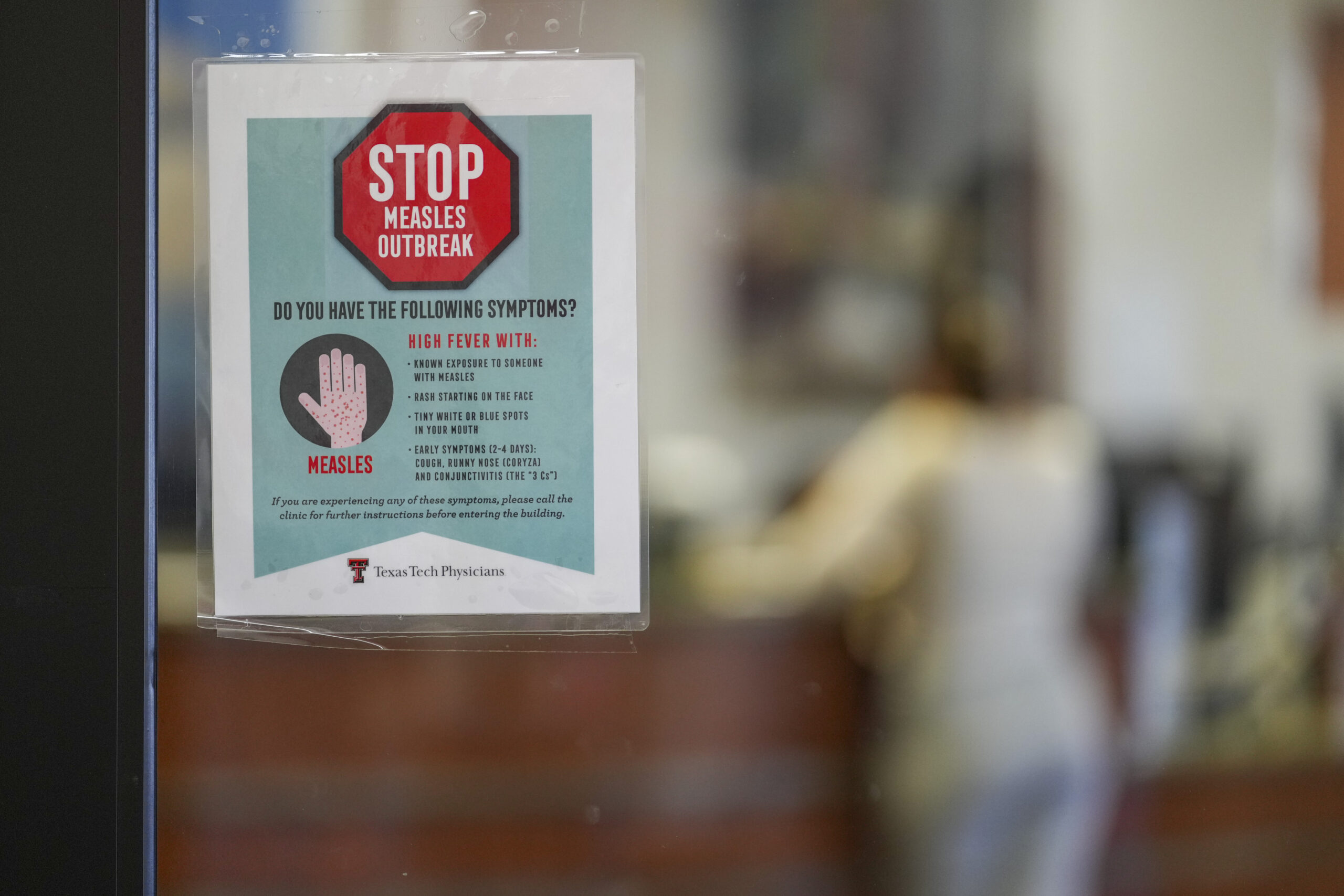 A sign on a glass door warns of a measles outbreak, listing symptoms like fever and rash. A person is blurred in the background, standing near a counter.