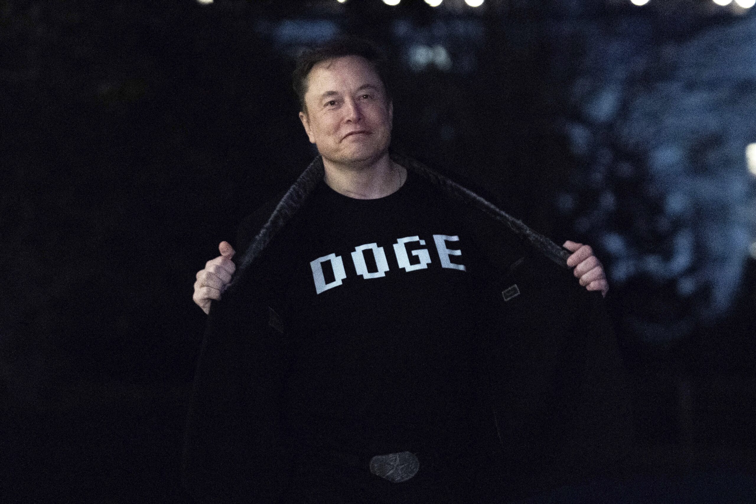 A person stands outdoors at night, opening their jacket to reveal a T-shirt with the word DOGE printed in bold letters.