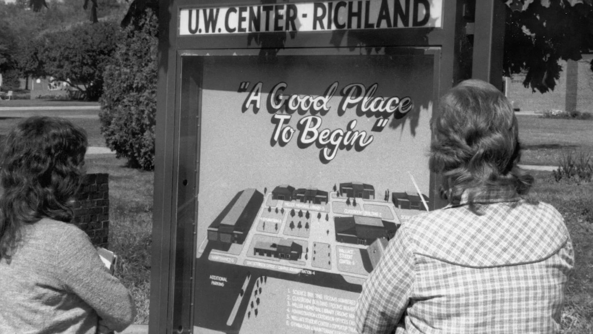 Two people stand in front of a sign for U.W. Center - Richland, reading A Good Place To Begin, with a map of the campus layout displayed.