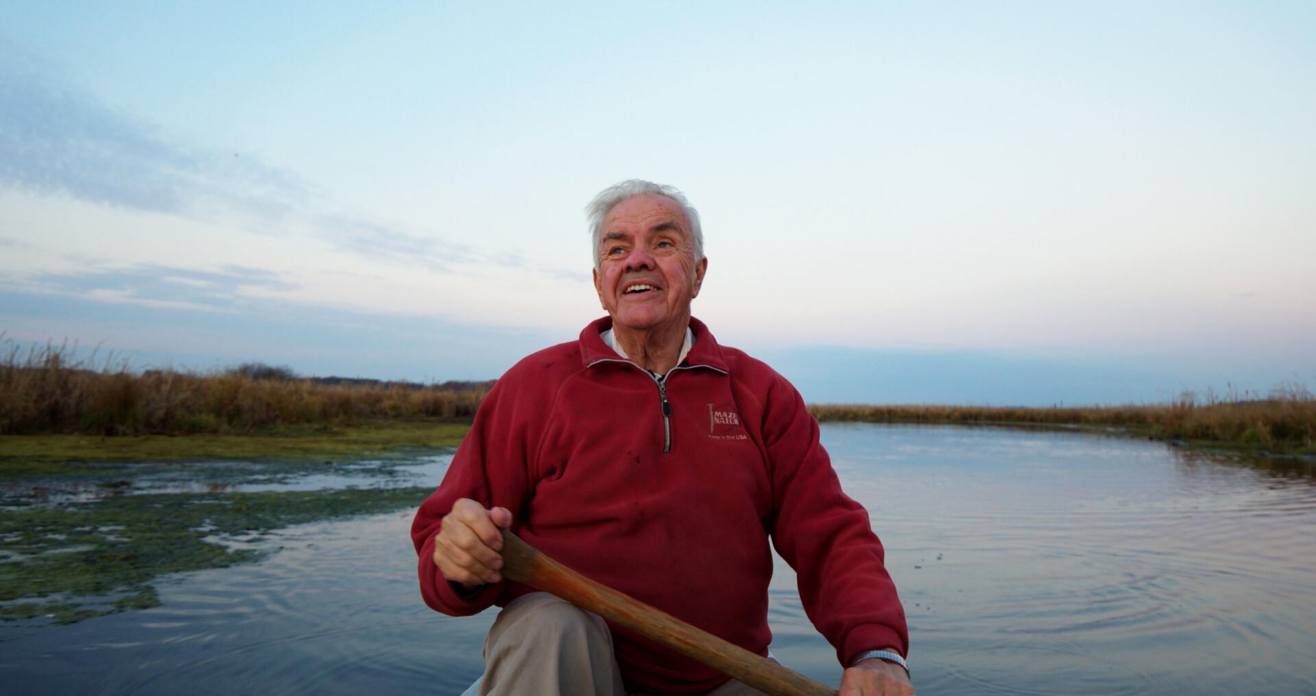 Older man in red sweater rowing a boat on a calm lake at dusk, with a clear sky and marshy shoreline in the background.