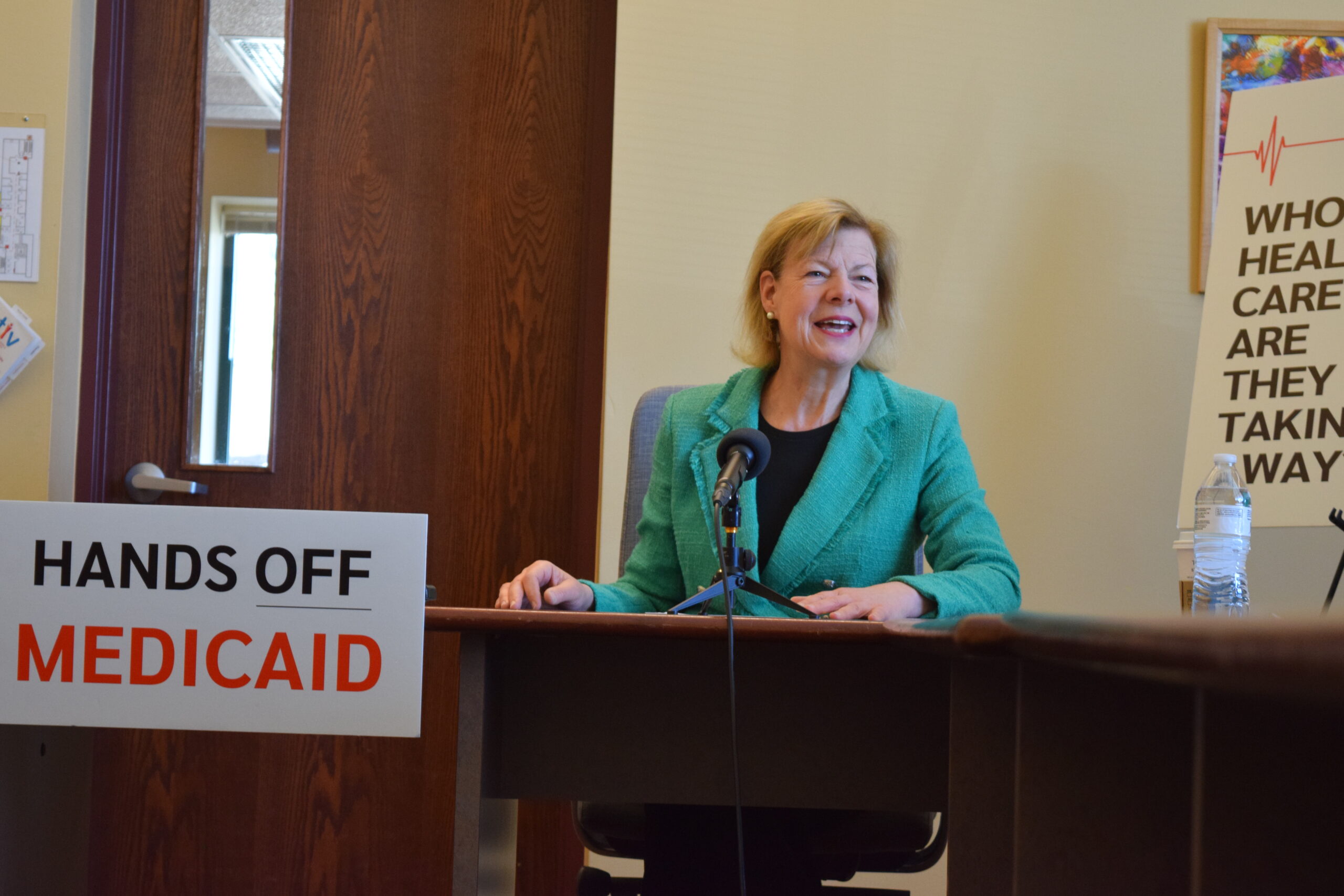 Person in a green blazer sits at a desk with a microphone, beside a sign reading HANDS OFF MEDICAID.