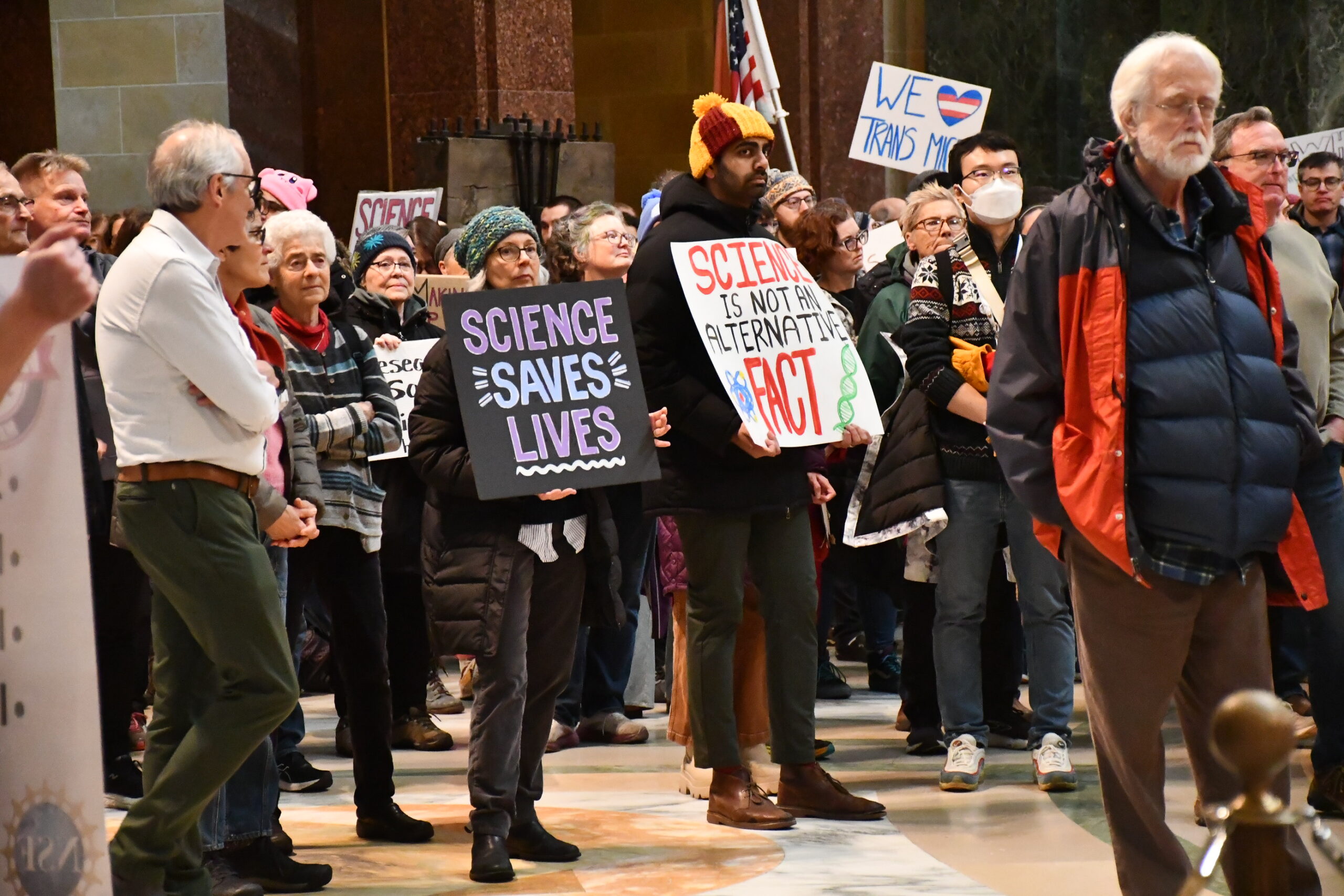 A group of people gathers indoors, holding signs that say Science Saves Lives and Science is Not an Alternative Fact.