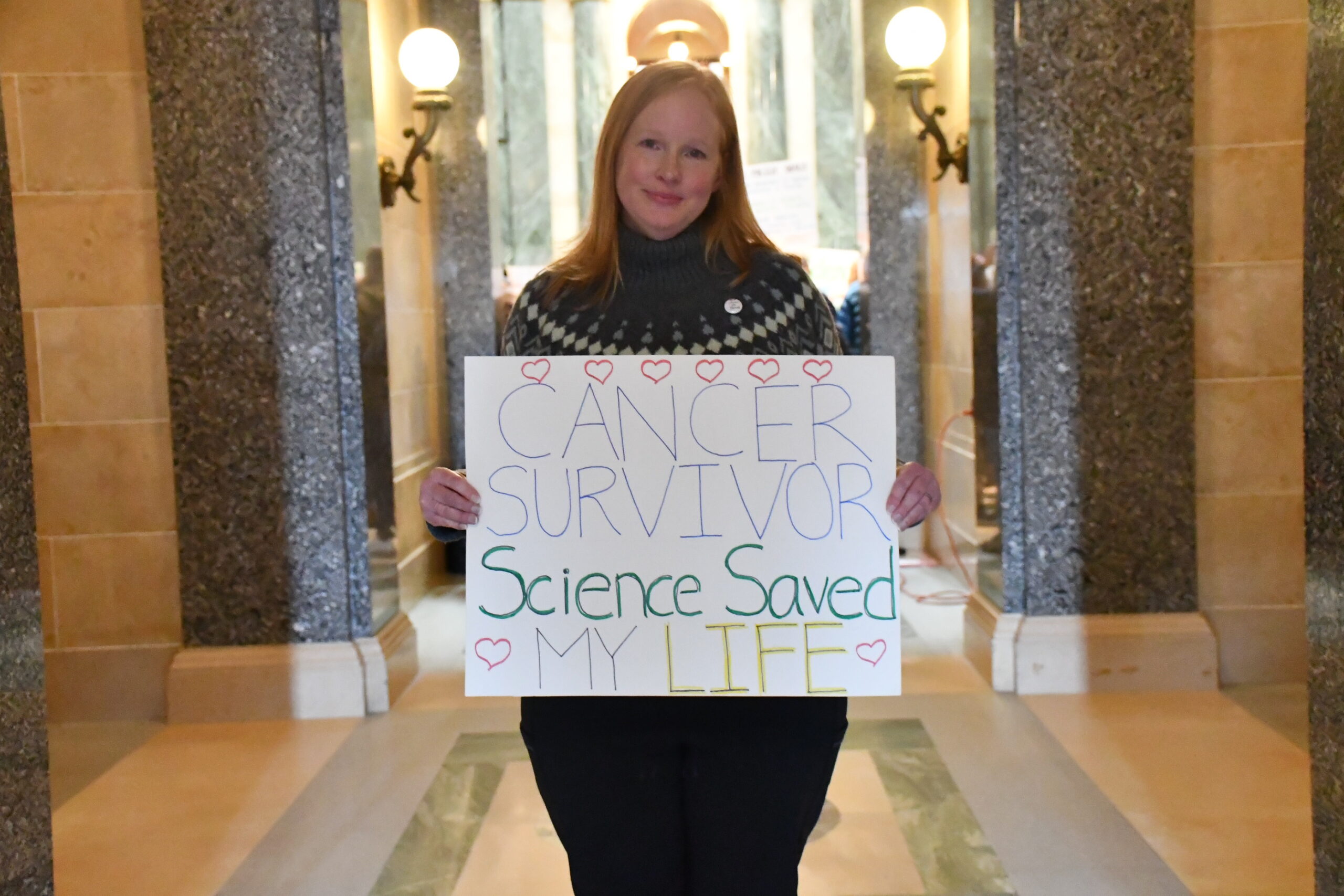 Person holding a sign that reads, Cancer Survivor, Science Saved My Life, standing in a building with ornate details.