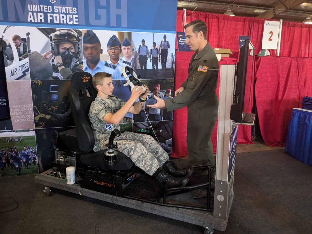 A young person in a military uniform sits in a flight simulator seat, holding a joystick, while another person in a military uniform stands nearby handing something to the seated individual.