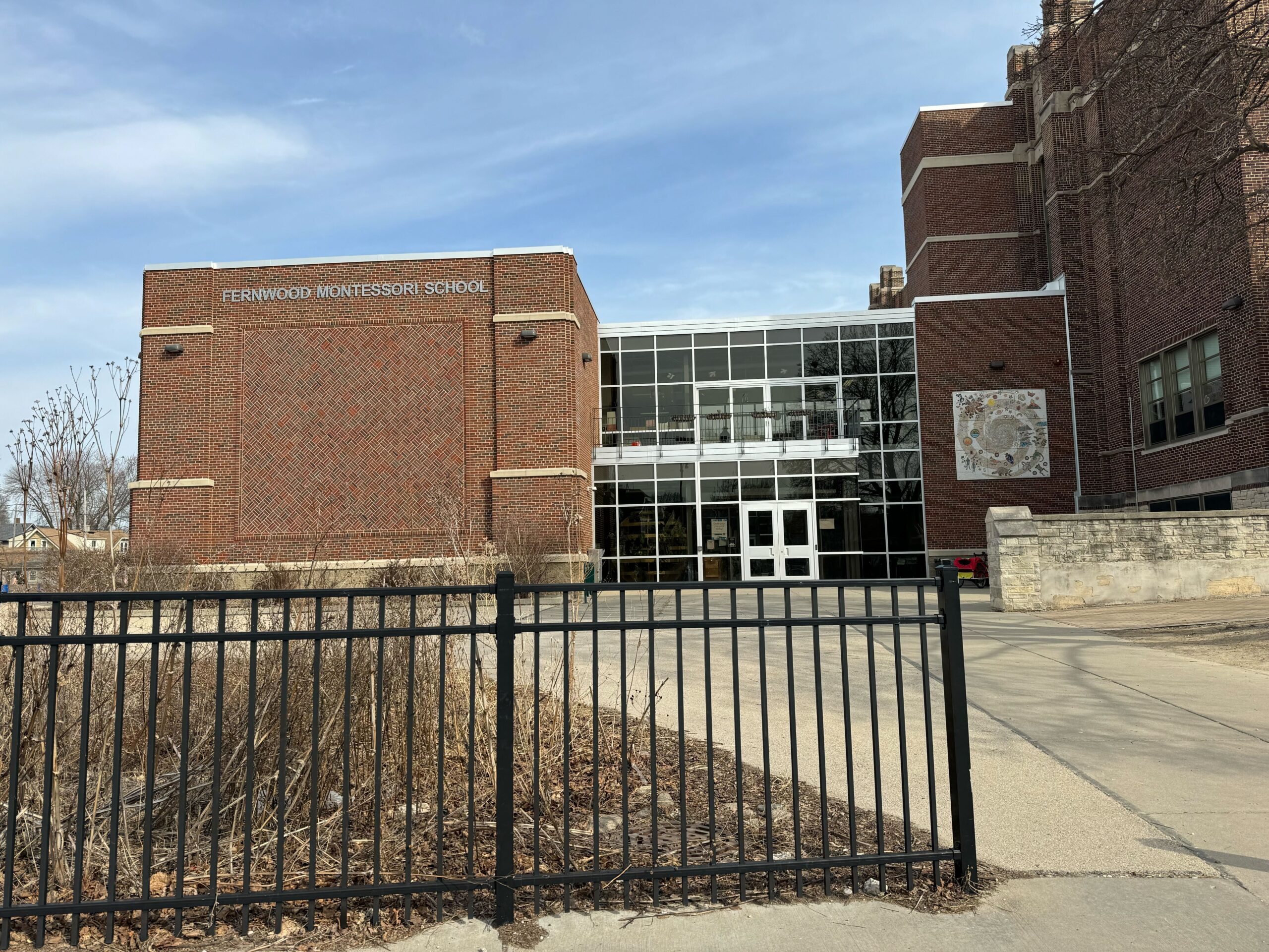 Brick school building with a sign reading Fernwood Montessori School. The entrance has glass doors and is surrounded by a black metal fence.