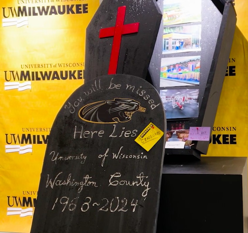 A tombstone display reads You will be missed, Here Lies University of Wisconsin Washington County, 1968-2024 with a refrigerator in the background and a red cross on the tombstone.