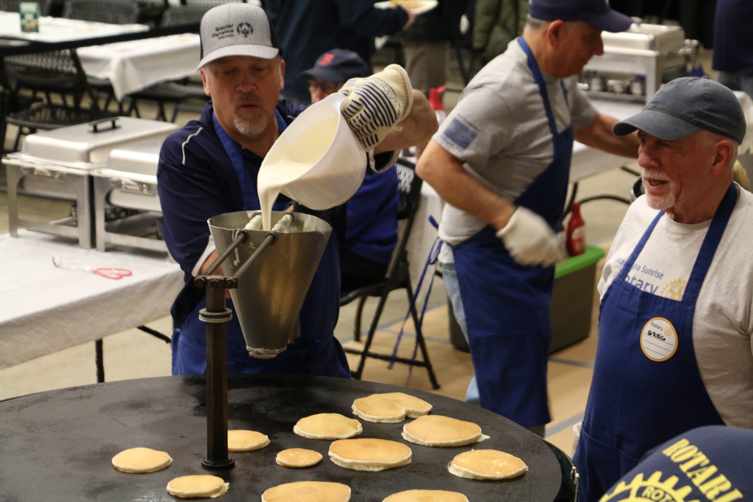 People making pancakes on a large griddle at an event; one pours batter while others prepare food, wearing uniforms and name tags.