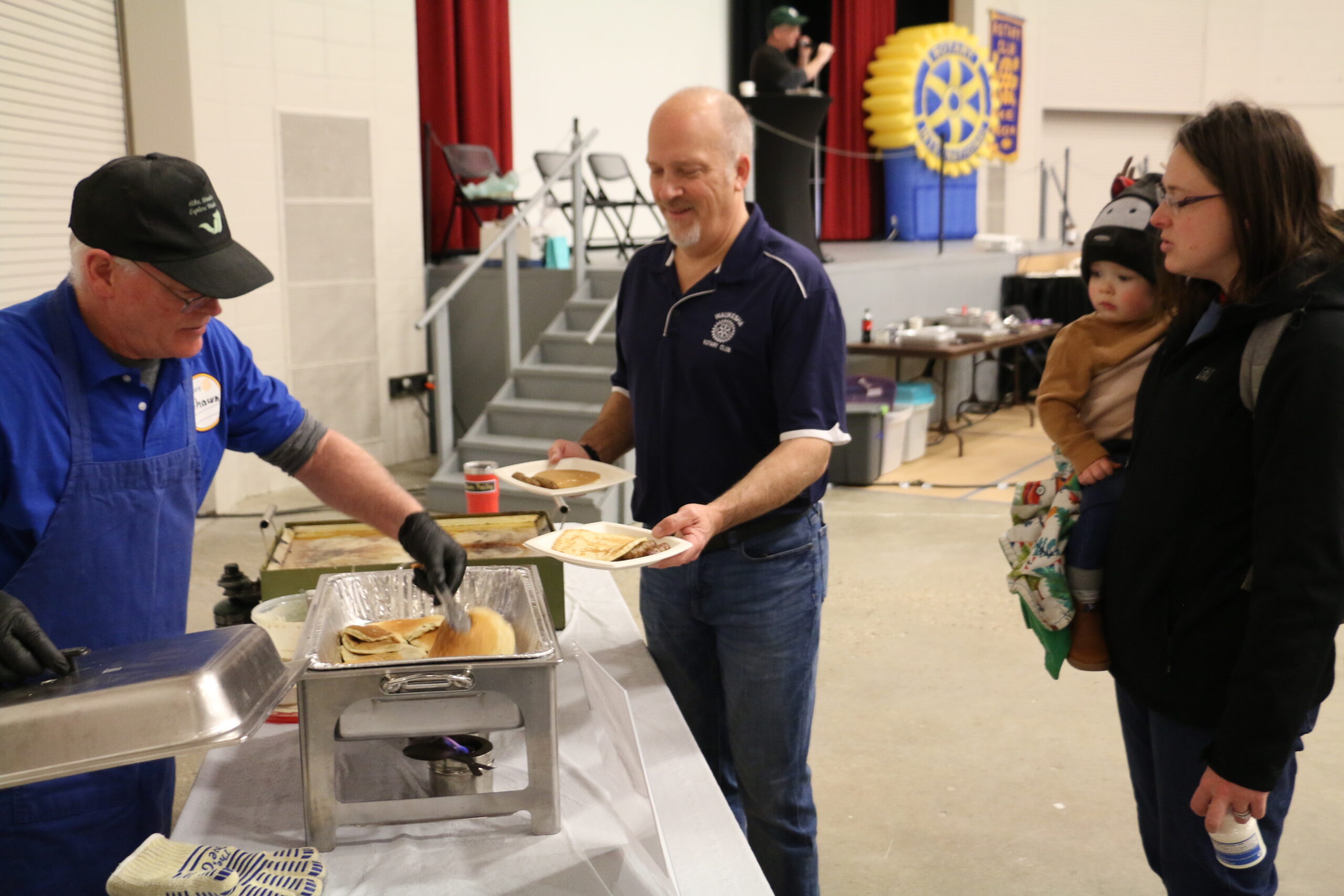 A person in a blue uniform serves pancakes to a man and a woman holding a child in a cafeteria setting.