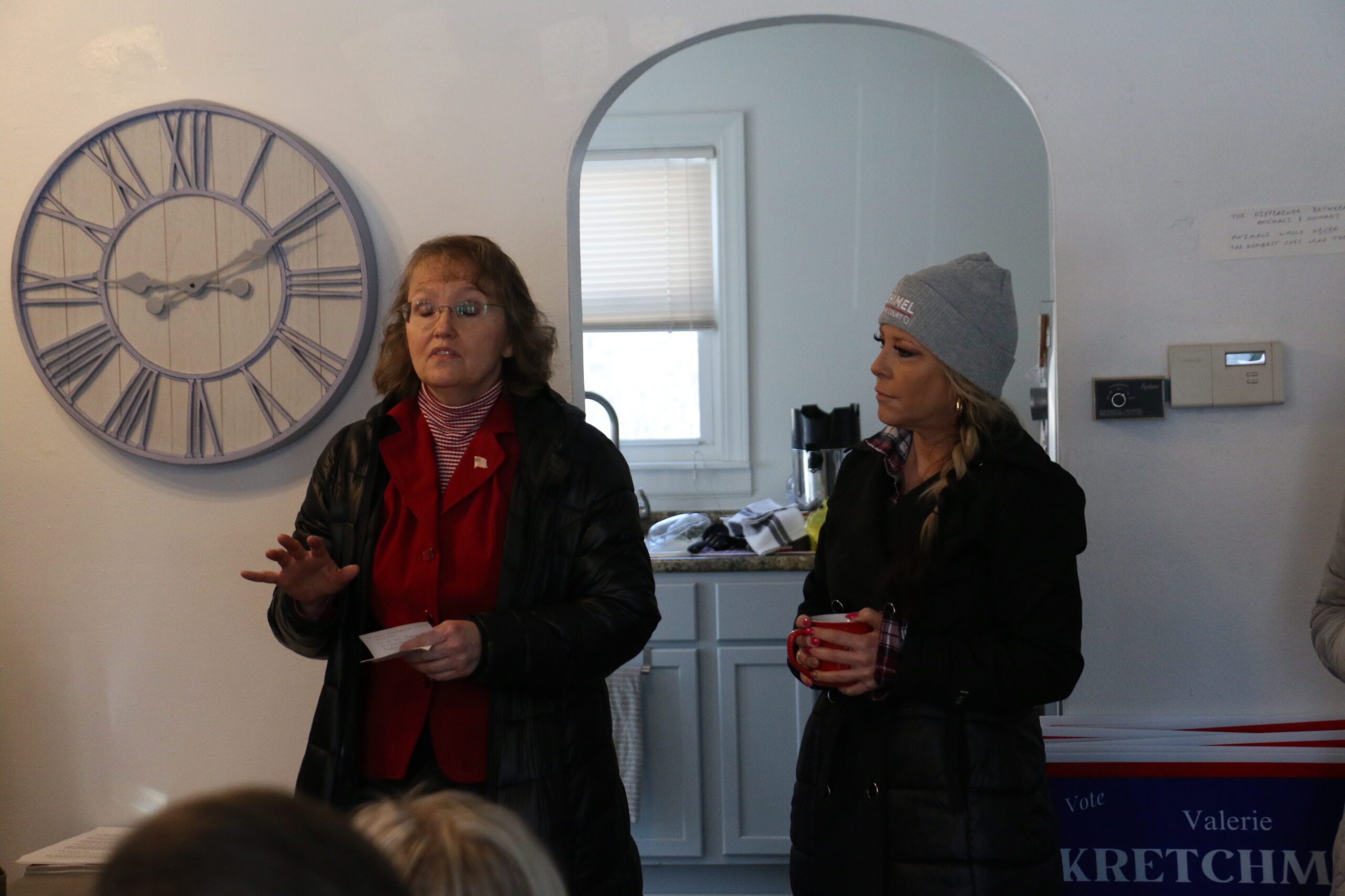 Two women stand in a room with a clock on the wall. One speaks while holding paper; the other holds a mug and wears a beanie. A sign with partial text is visible on the right.