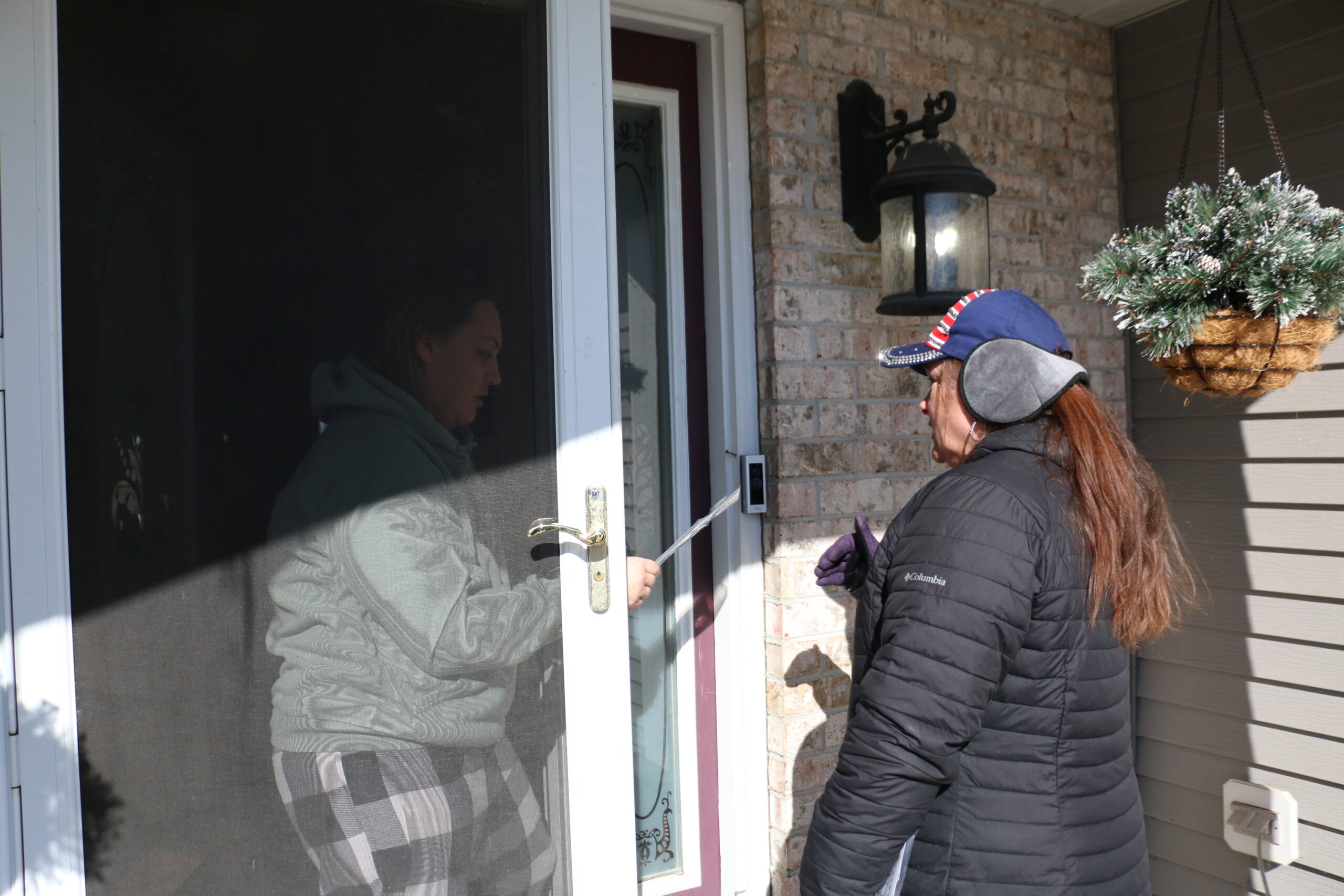 Person standing outside a storm door, holding out a pen; another person is visible behind the door, observing. A hanging planter and outdoor light fixture are on the brick wall.