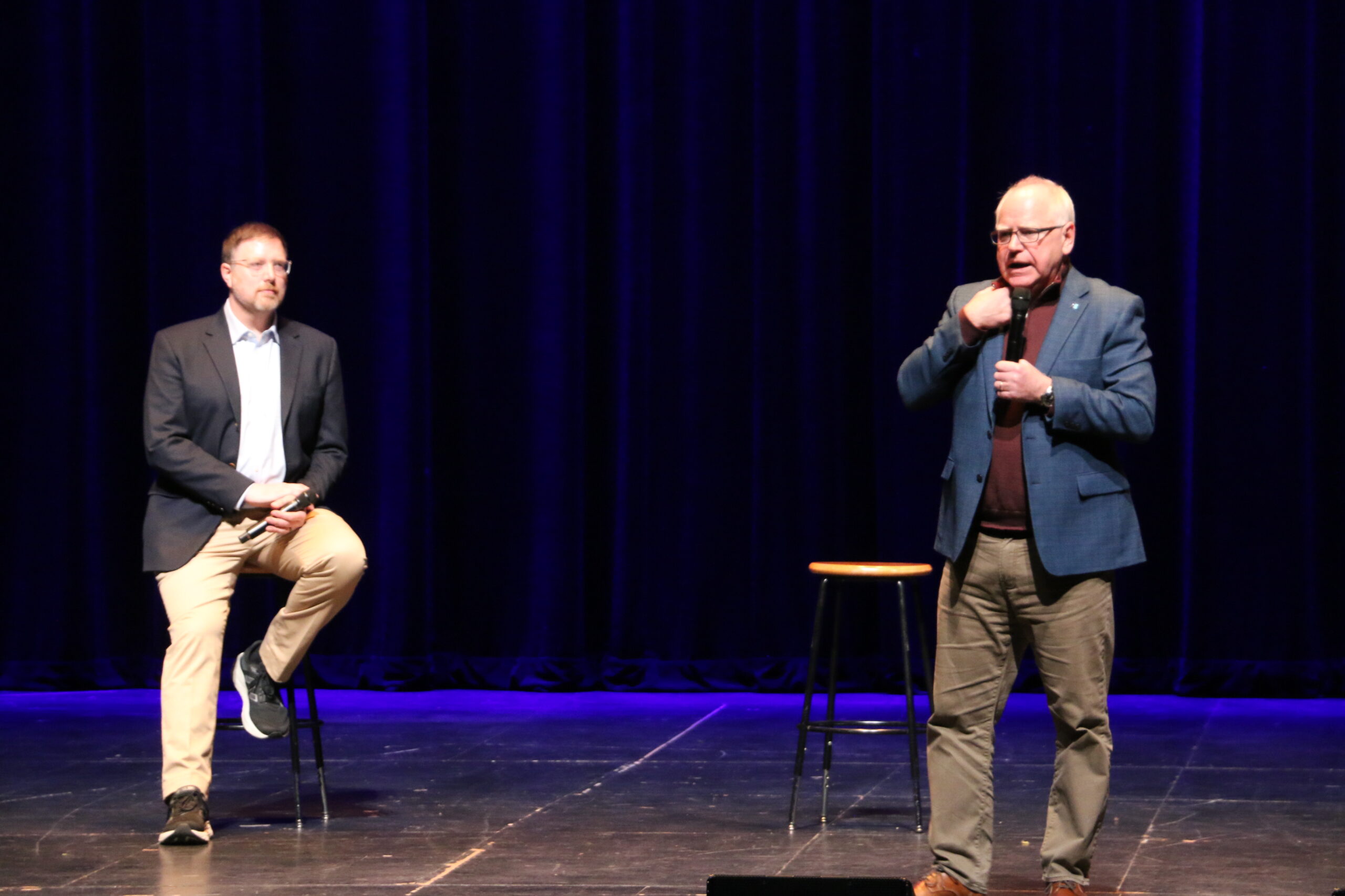 Two men are on stage with a dark blue curtain backdrop. One man stands and speaks into a microphone, gesturing with his hand, while the other man sits on a stool, listening attentively.