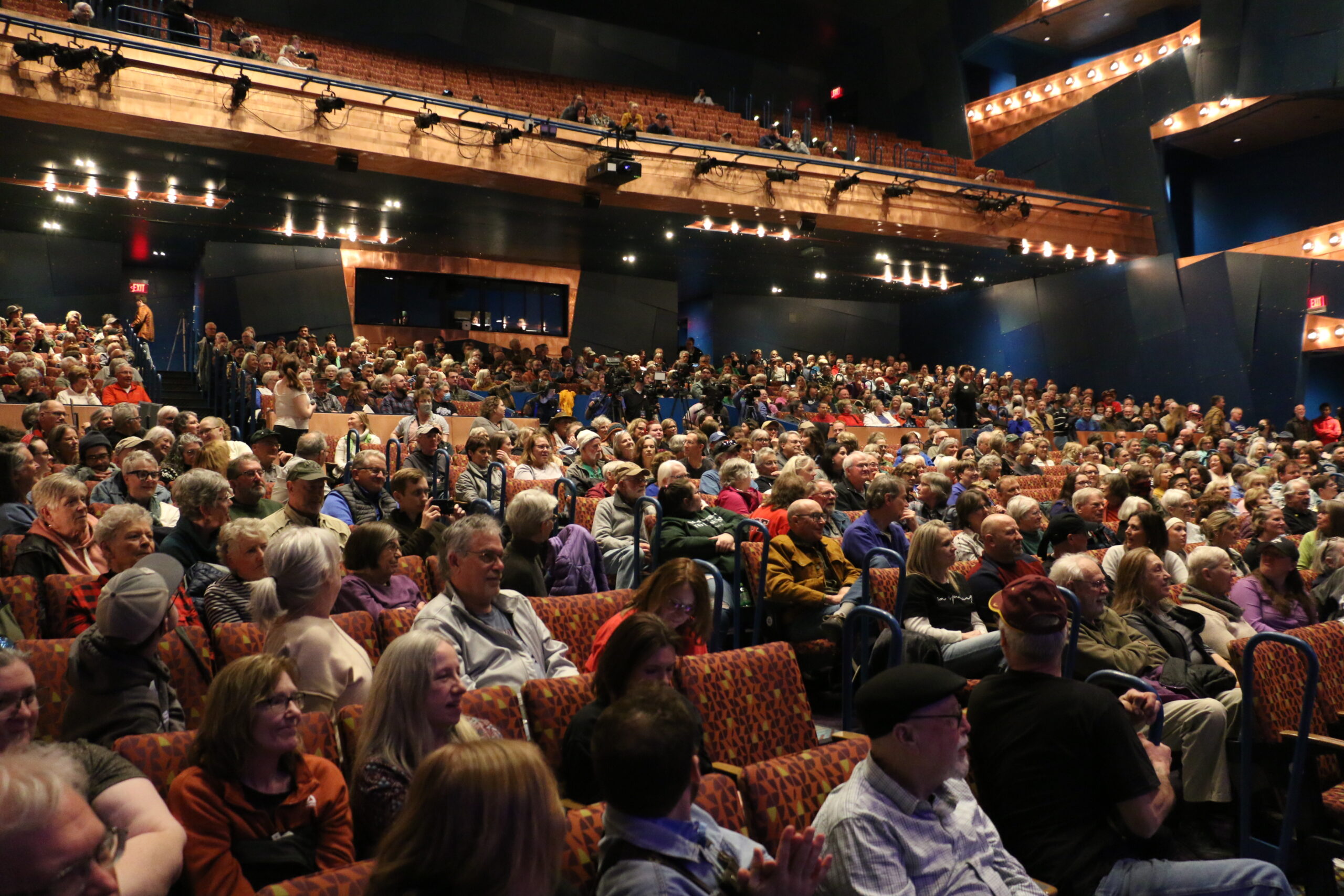 A large audience seated in a theater with rows of red upholstered chairs, attentively watching a performance on stage.