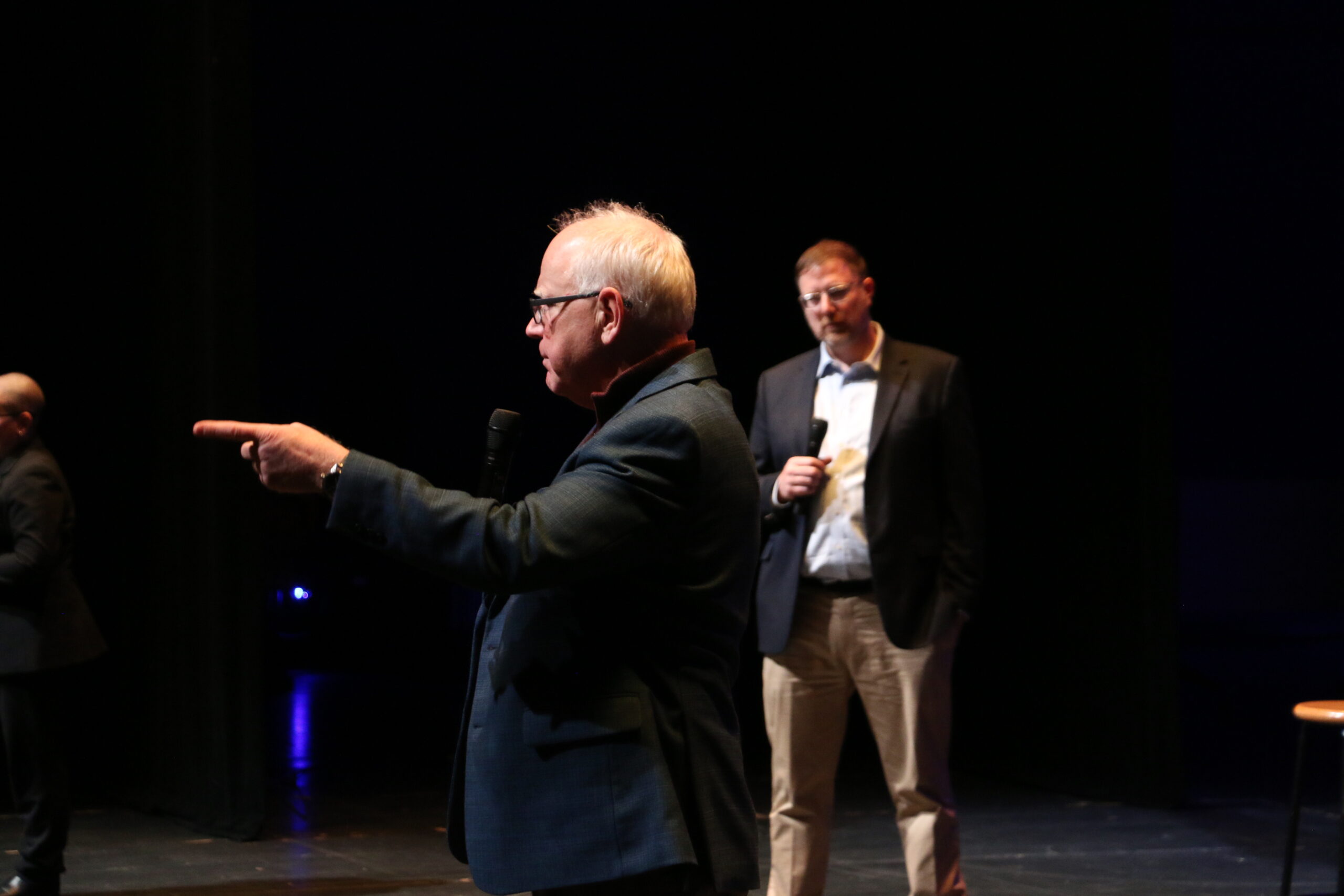 Two men in suits stand on a dimly lit stage. The man in the foreground points while speaking into a microphone, and the man in the background holds a microphone by his side.