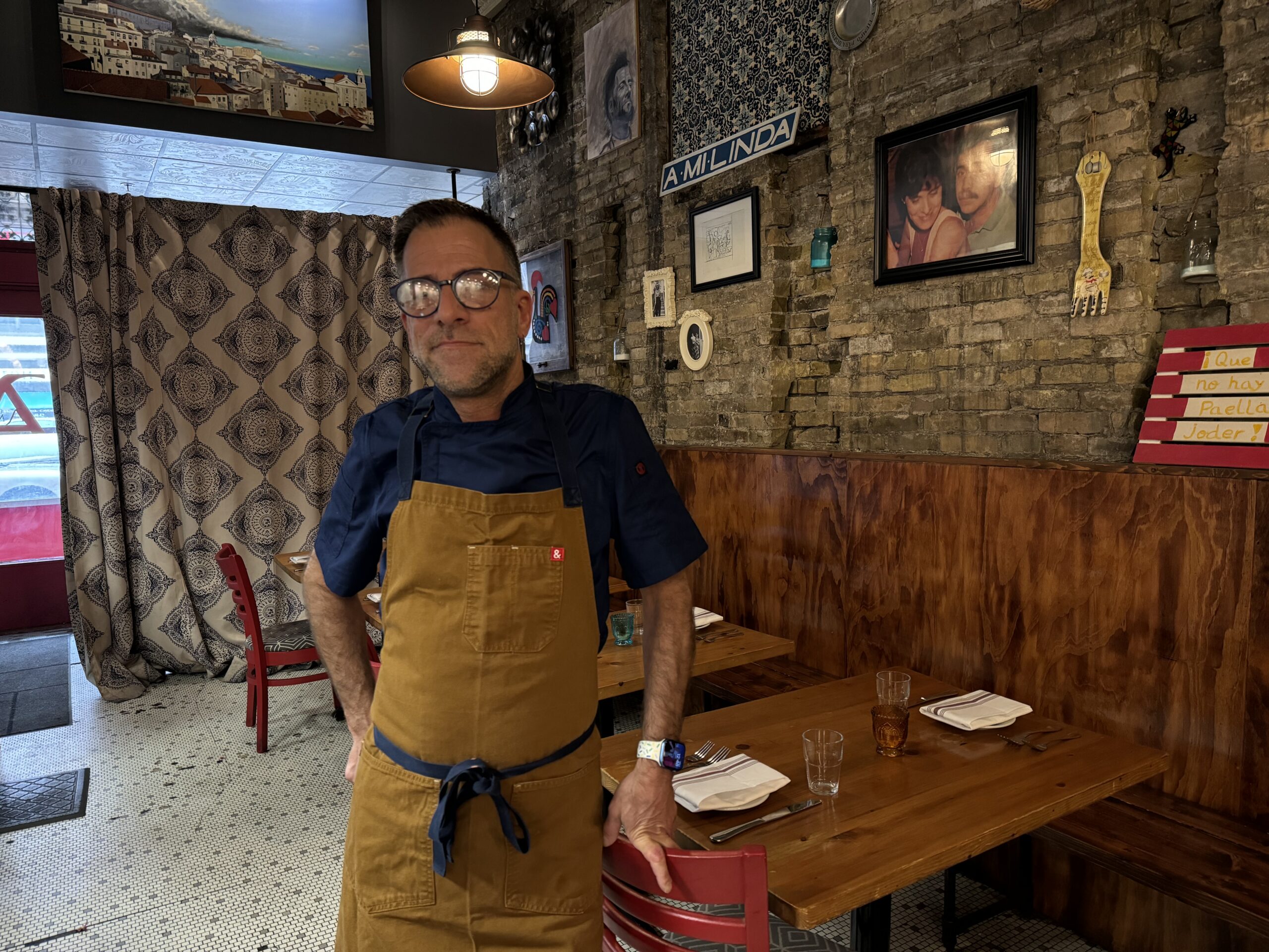 A man in glasses and an apron stands inside a restaurant with rustic brick walls and wooden tables set with plates.